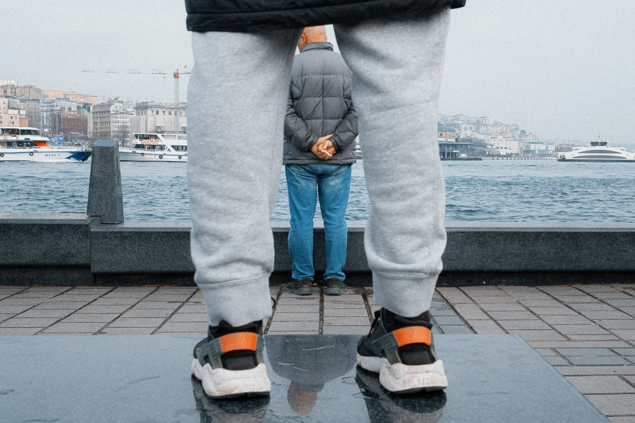 Une personne est vue à travers une grande paire de chaussures et un pantalon gris, regardant un homme en veste grise bord de l'eau avec des bateaux en arrière-plan.