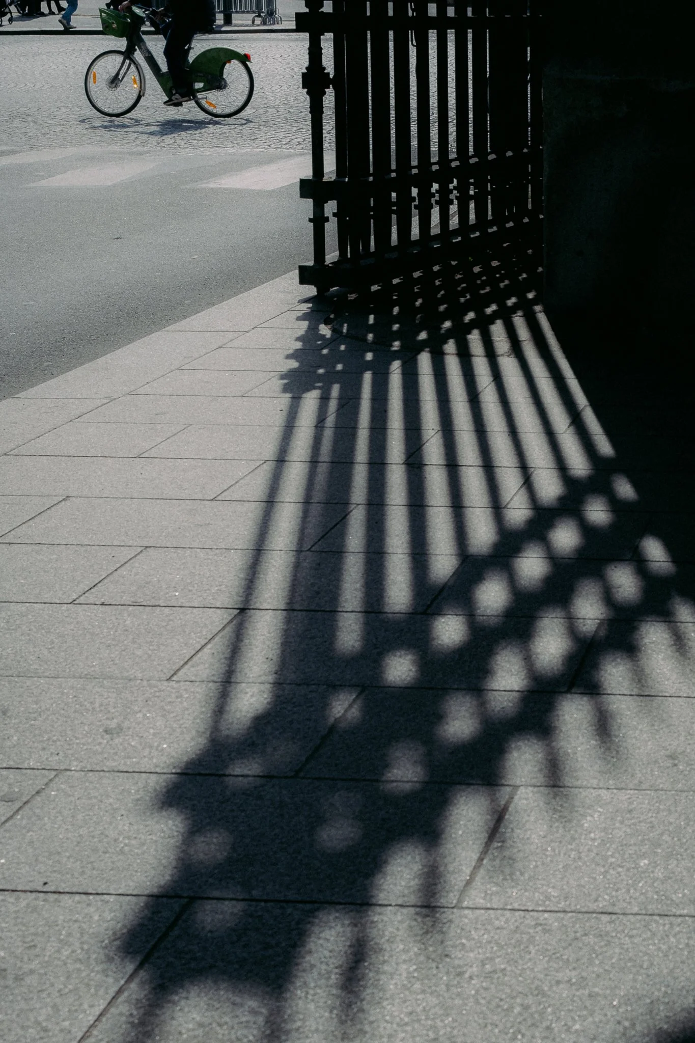 Ombre d'une grille en métal sur le trottoir, avec un vélo vert passant au loin, dans une rue pavée lors d'une journée ensoleillée.