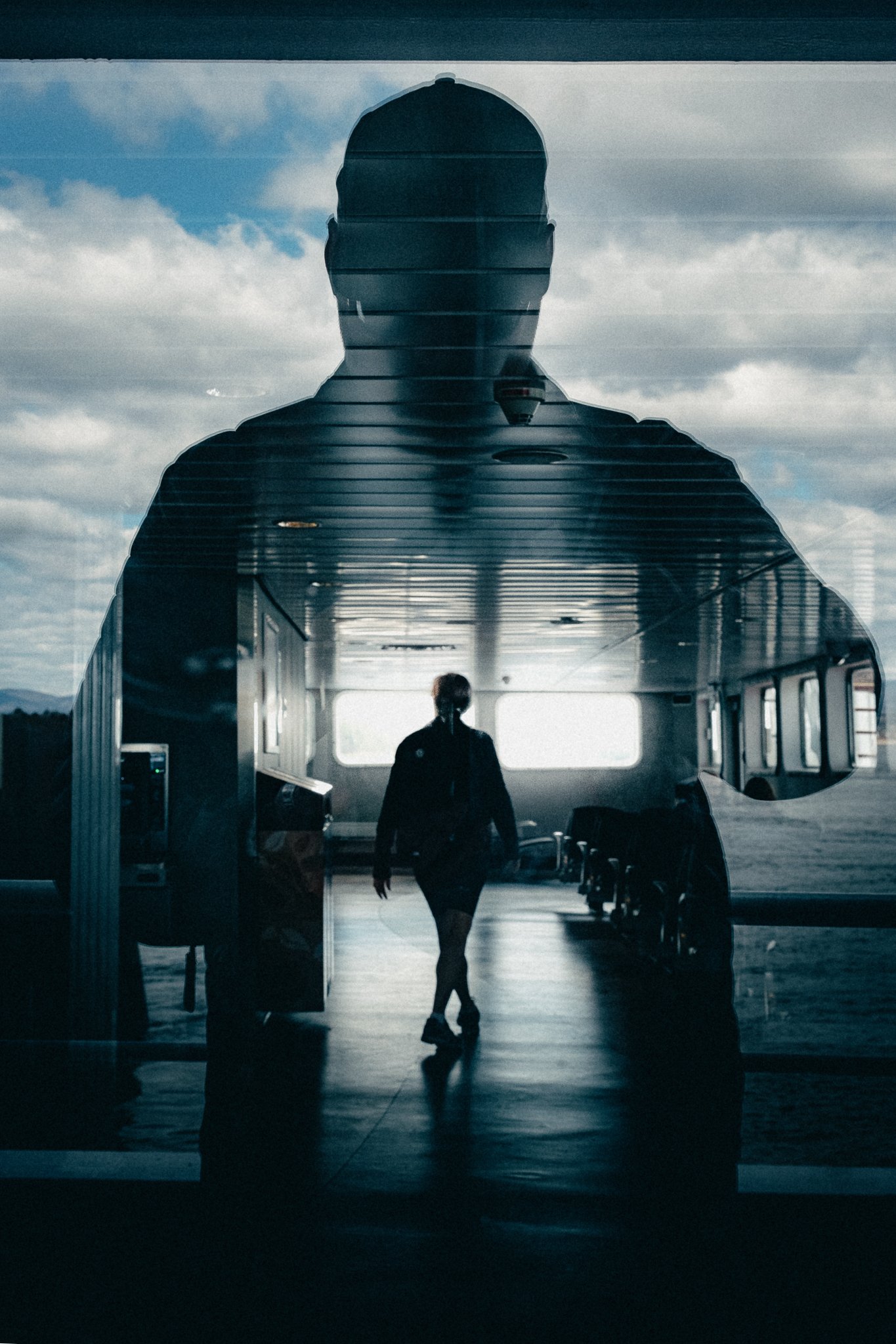 Une personne marche à travers une porte d'aéroport ou de station de train, vue de l'intérieur, avec des grandes fenêtres et un ciel nuageux visible à l'extérieur.