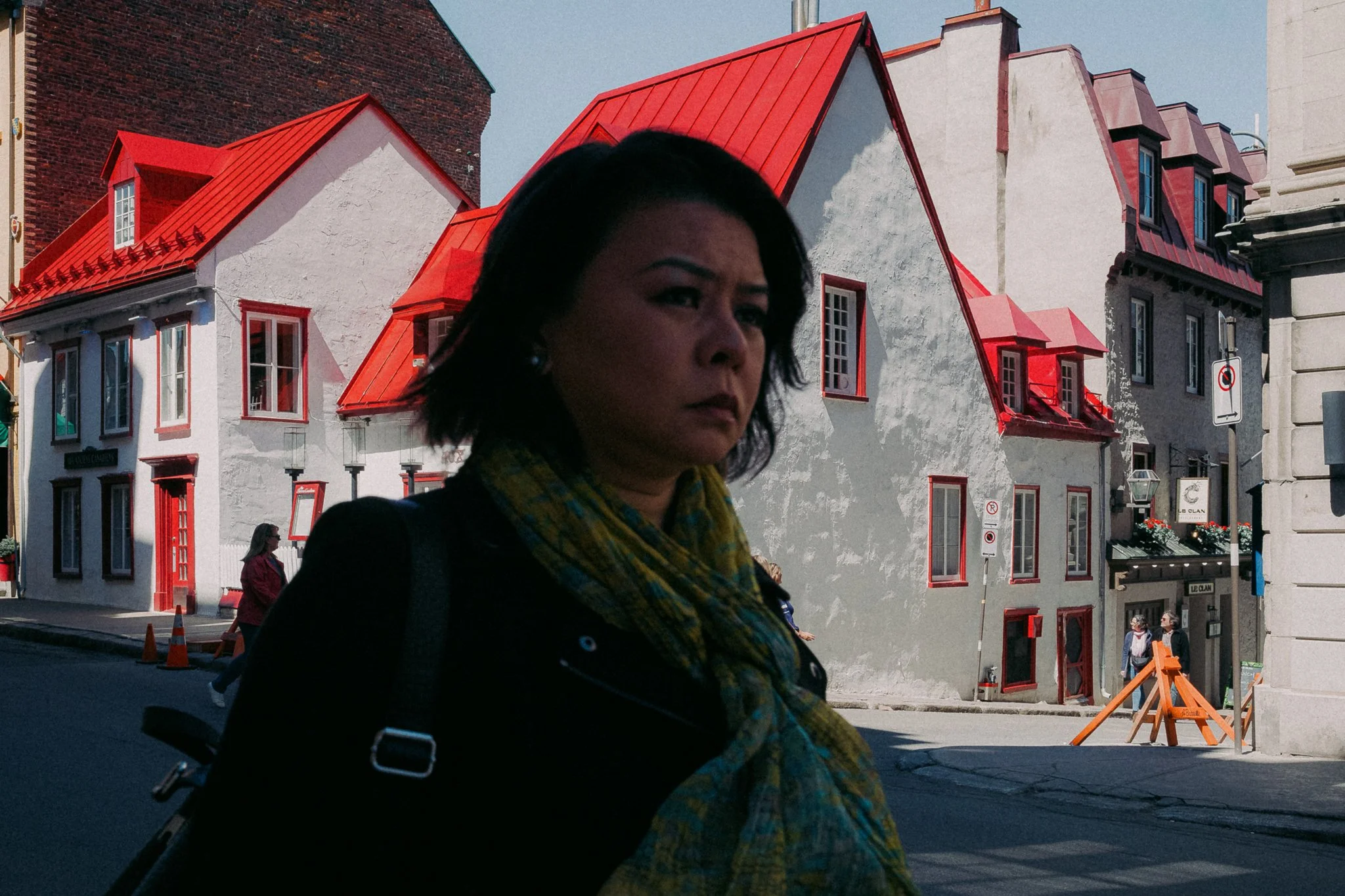 Une femme avec un foulard coloré marche dans une rue urbaine avec des bâtiments en arrière-plan, dont une maison avec un toit rouge vif.