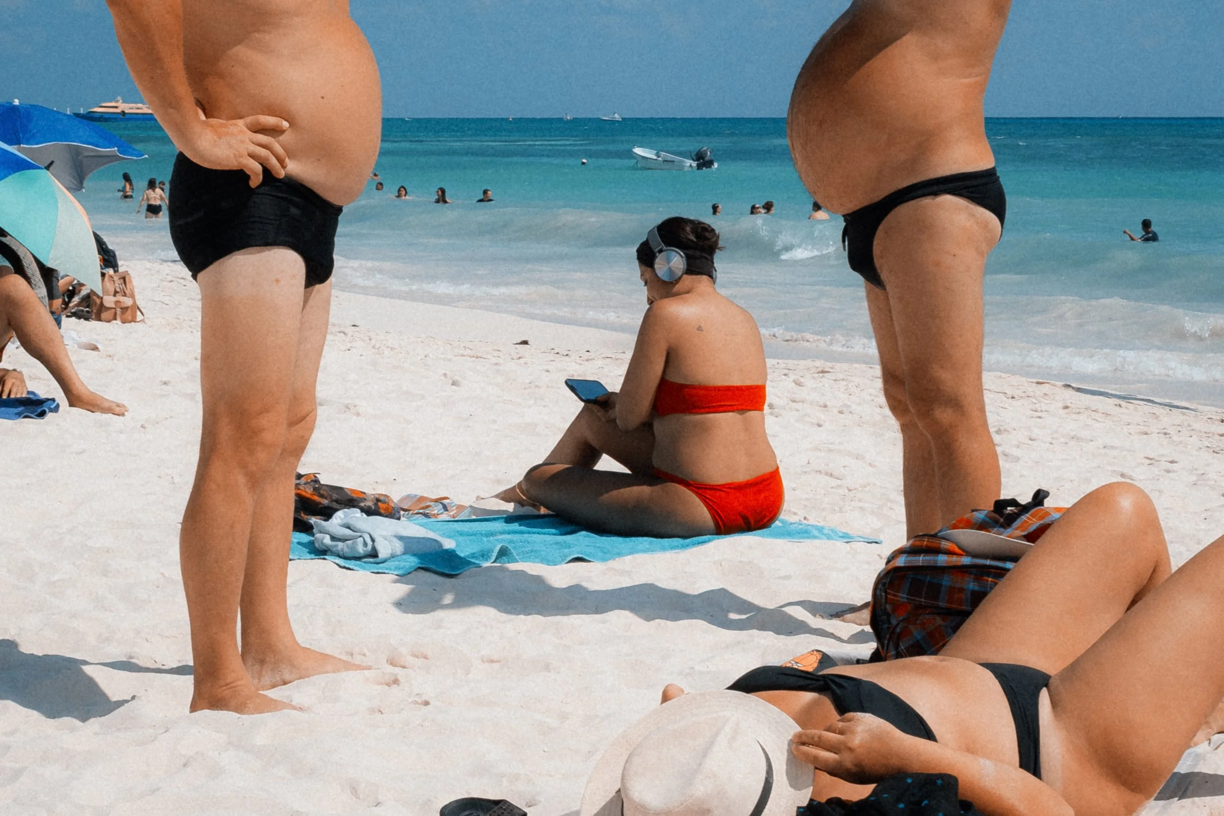 Vue de plage avec plusieurs personnes, deux adultes en maillots de bain noirs et un enfant en maillot rouge, assis ou debout sur le sable blanc. La mer turquoise en arrière-plan avec des bateaux et des parasols.