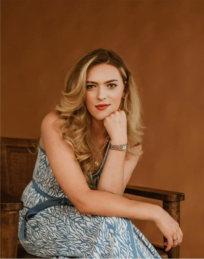 Woman with long wavy blonde hair wearing a sleeveless blue and white patterned dress, sitting on a wooden chair with a warm brown background.