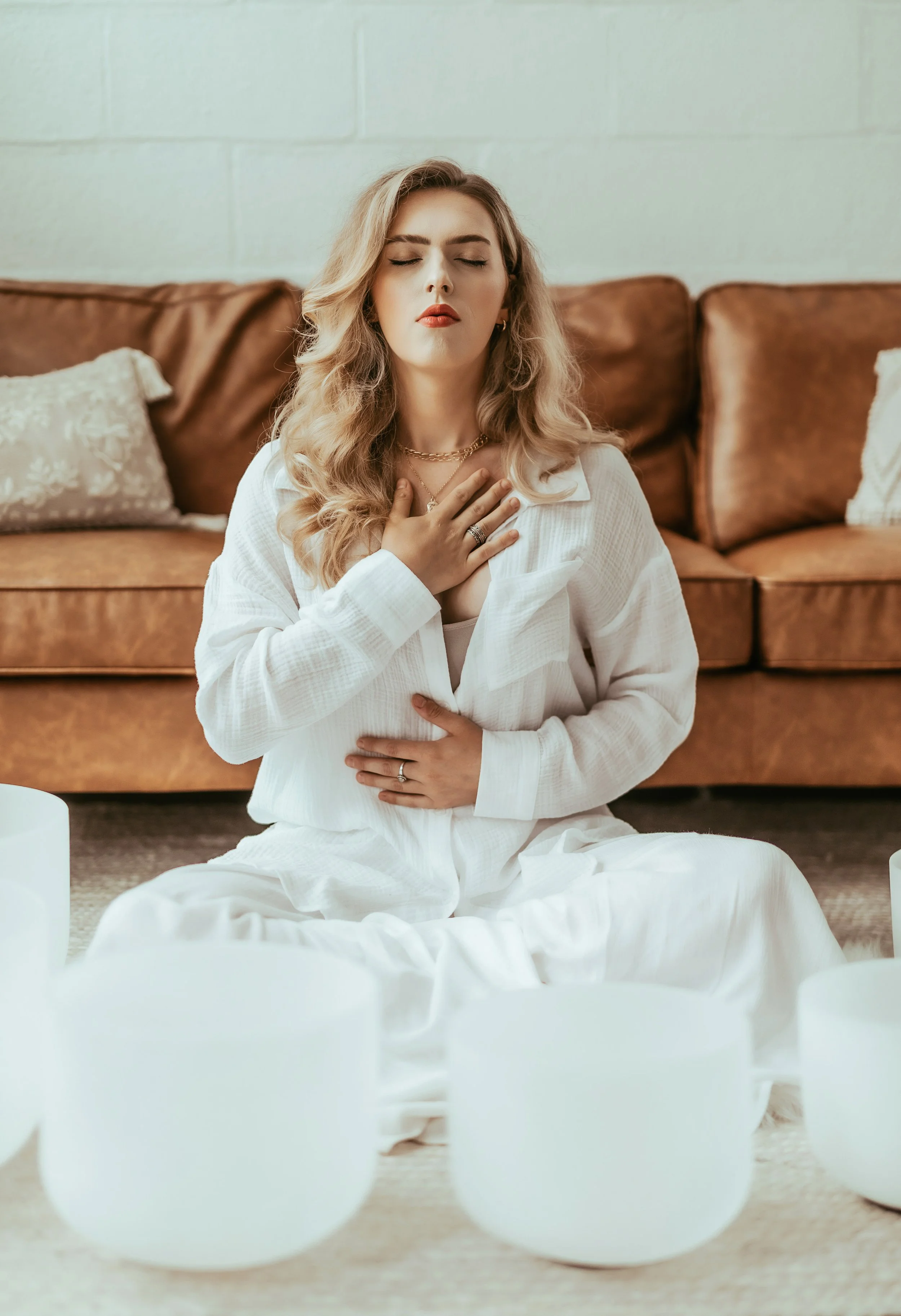 A woman with blonde, wavy hair and closed eyes meditating, sitting cross-legged on the floor with her right hand on her chest and her left hand on her stomach in front of white candles, a brown couch, and decorative pillows.