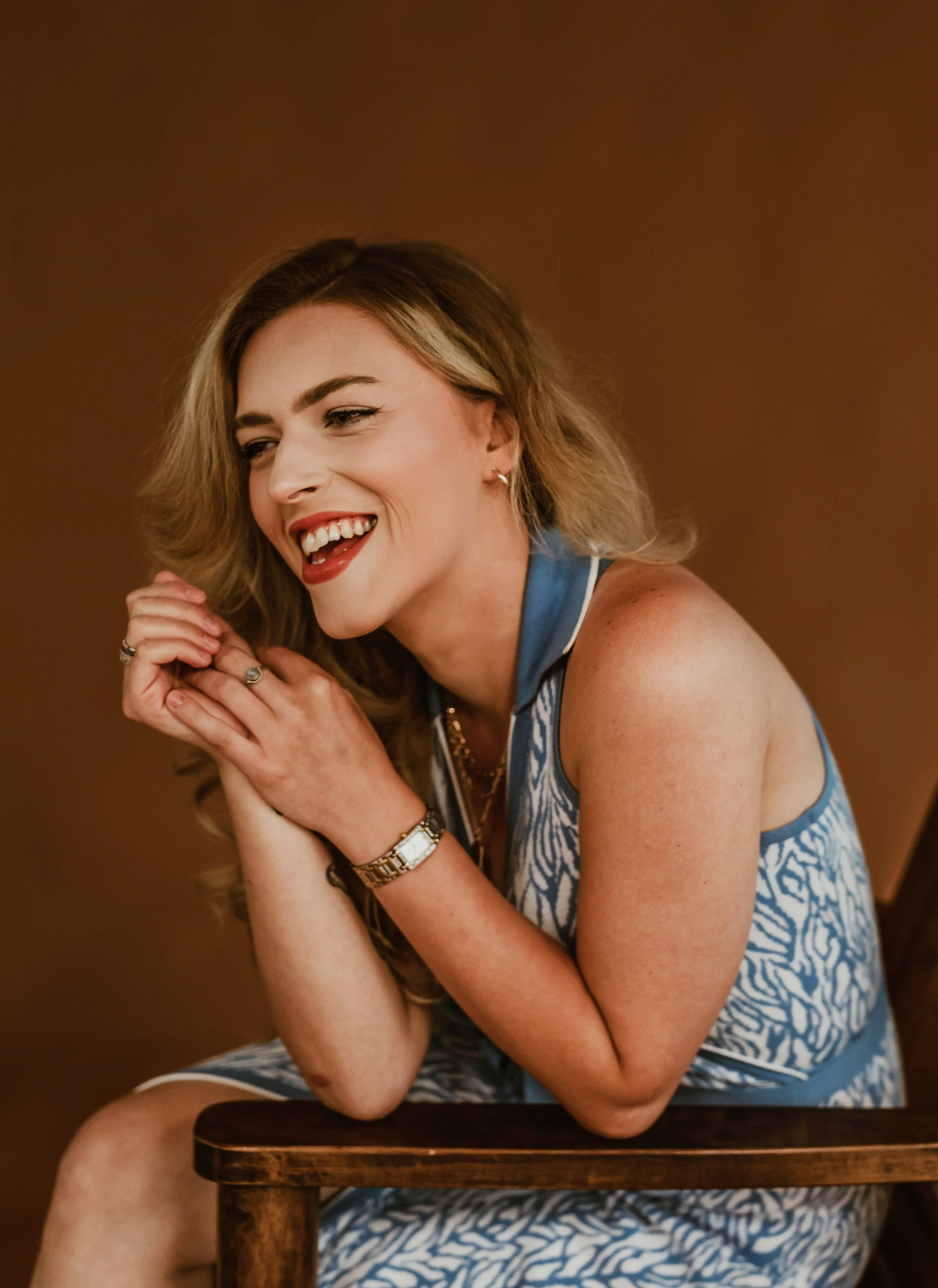 A woman with long wavy hair sitting and smiling, wearing a blue and white patterned sleeveless dress and jewelry, with her hands clasped together.