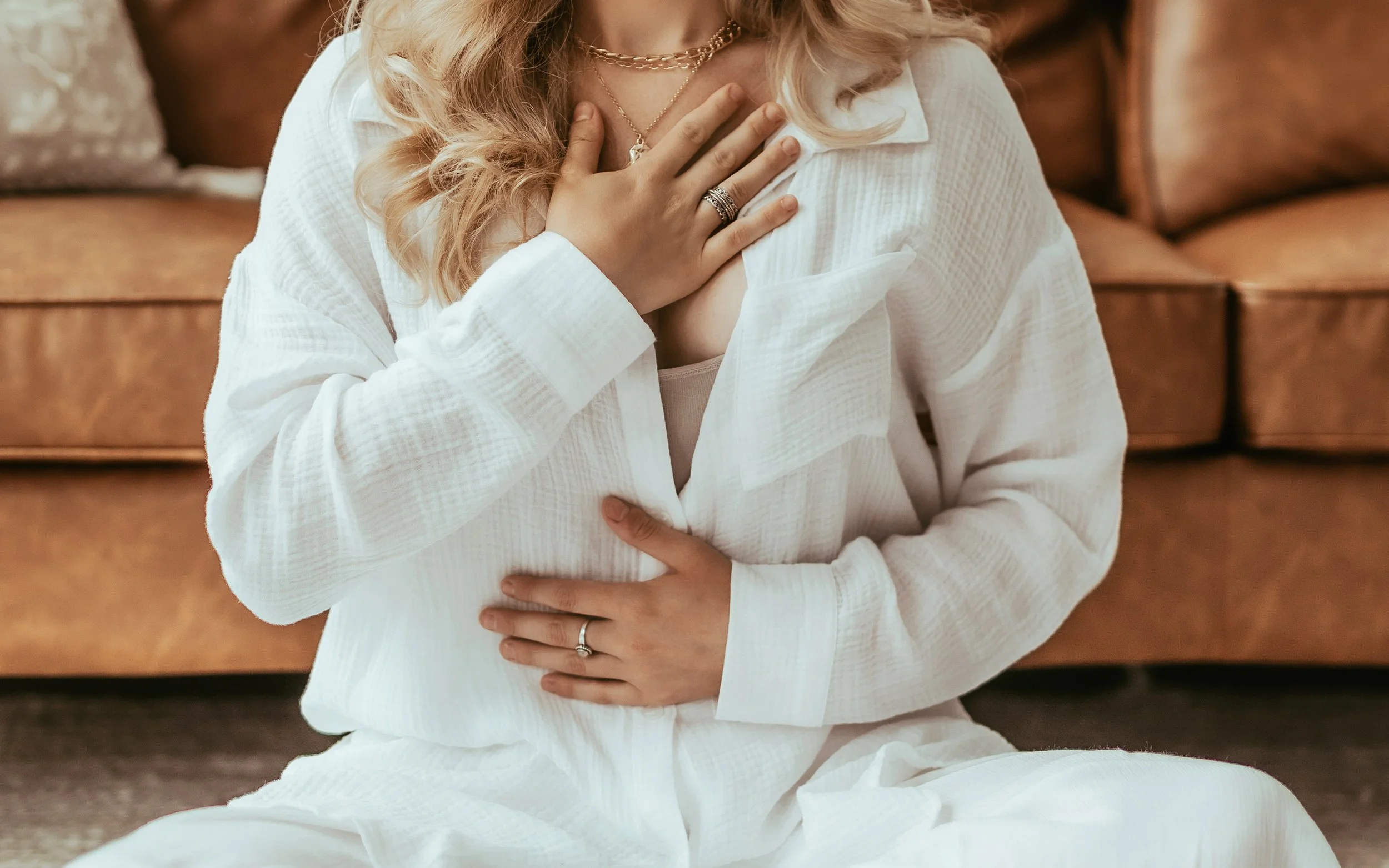 A woman sitting on the floor in front of a brown leather couch, wearing a white outfit, with her hand on her chest and stomach.