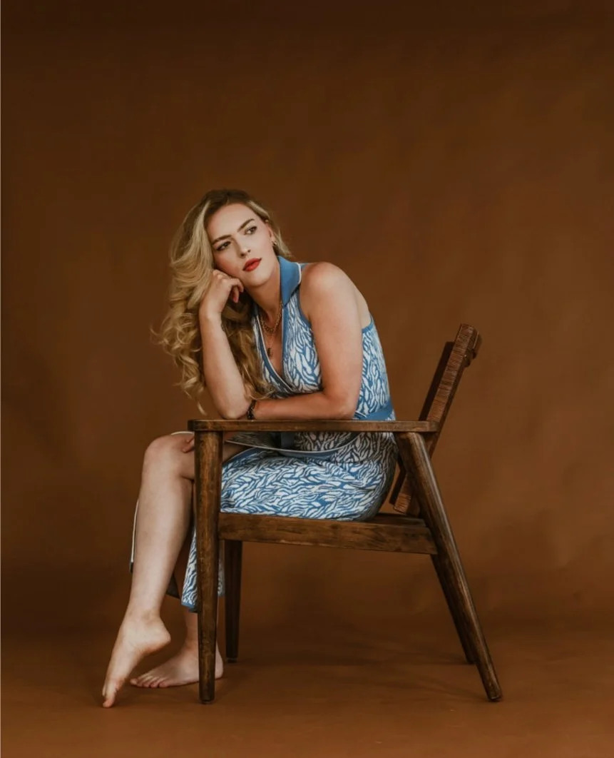 Woman with long blonde hair sitting on a wooden chair against a brown background, wearing a blue patterned dress.