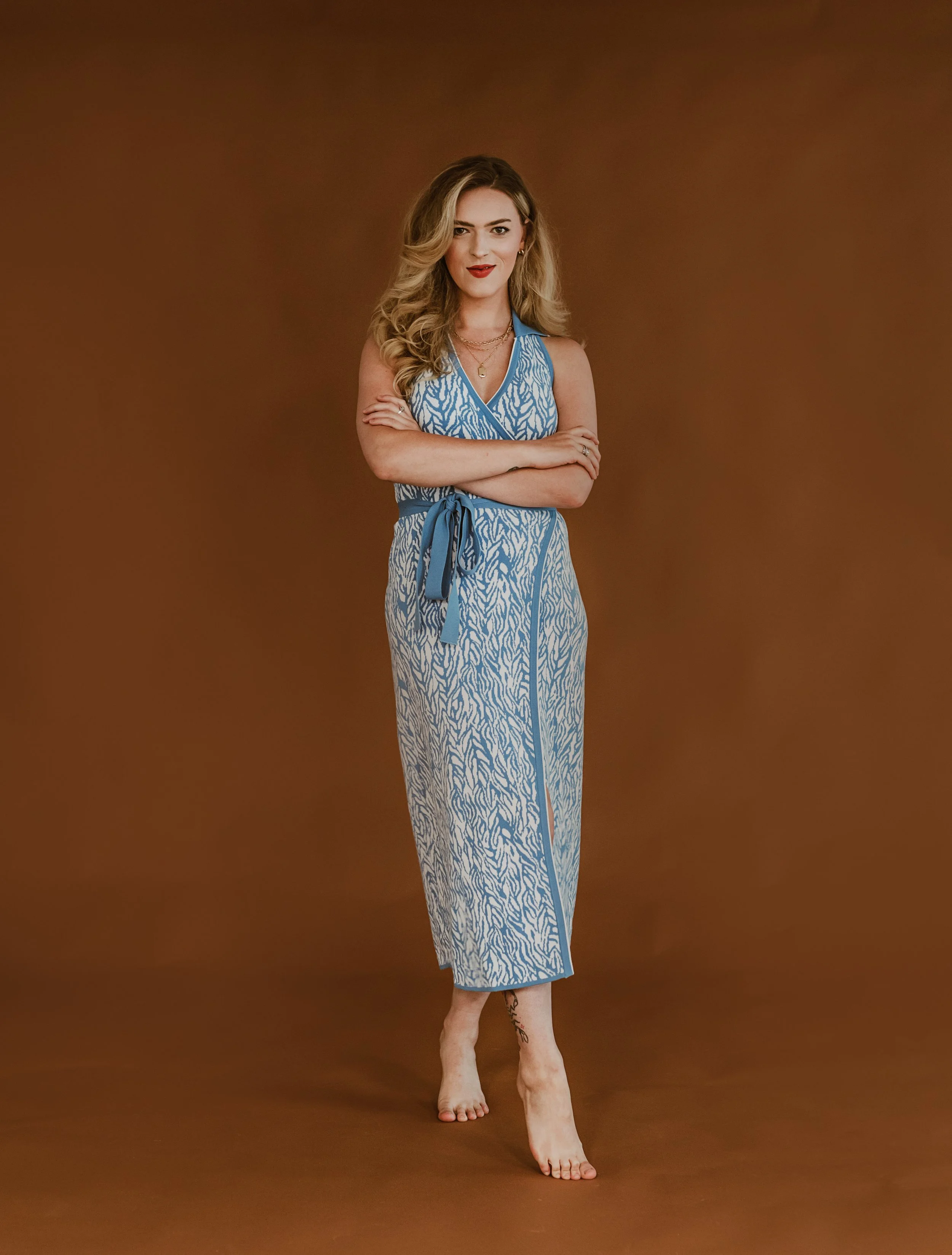 Woman with blonde hair, wearing a blue patterned sleeveless jumpsuit with a tie waist, standing barefoot against a brown background.