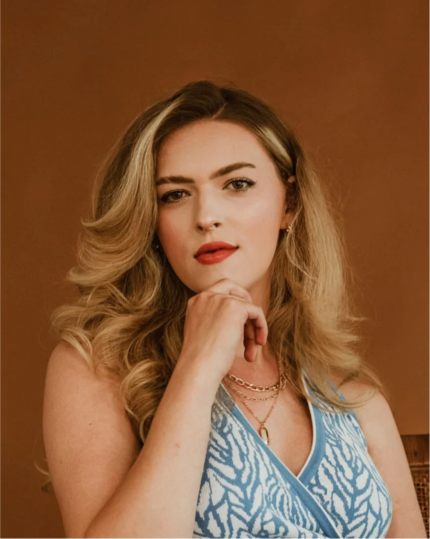 A woman with long wavy blonde hair, wearing a blue patterned sleeveless top, red lipstick, and layered gold necklaces, resting her chin on her hand against a plain brown background.
