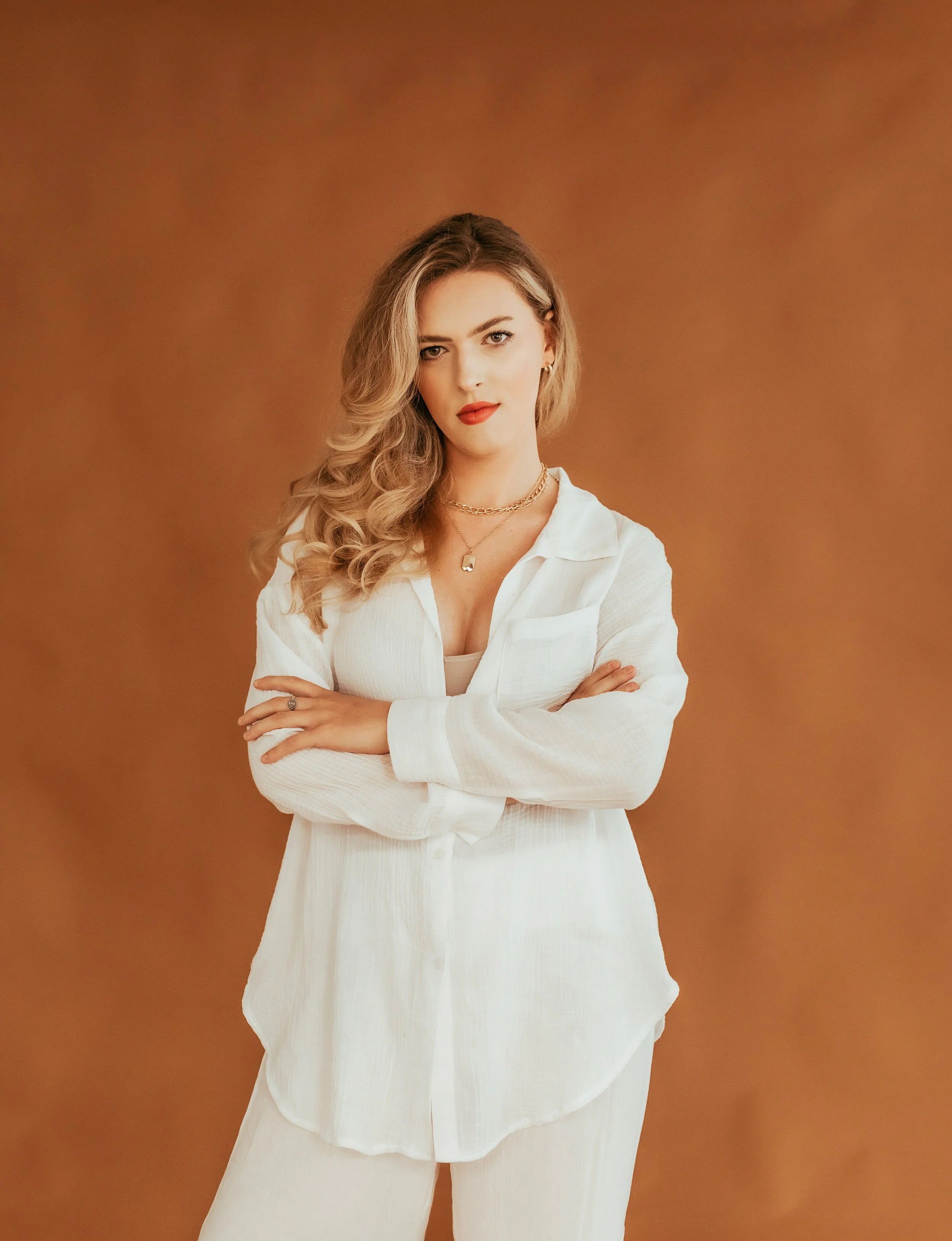 A woman with long blonde hair, wearing a white button-up shirt and white pants, standing with arms crossed against a brown background.
