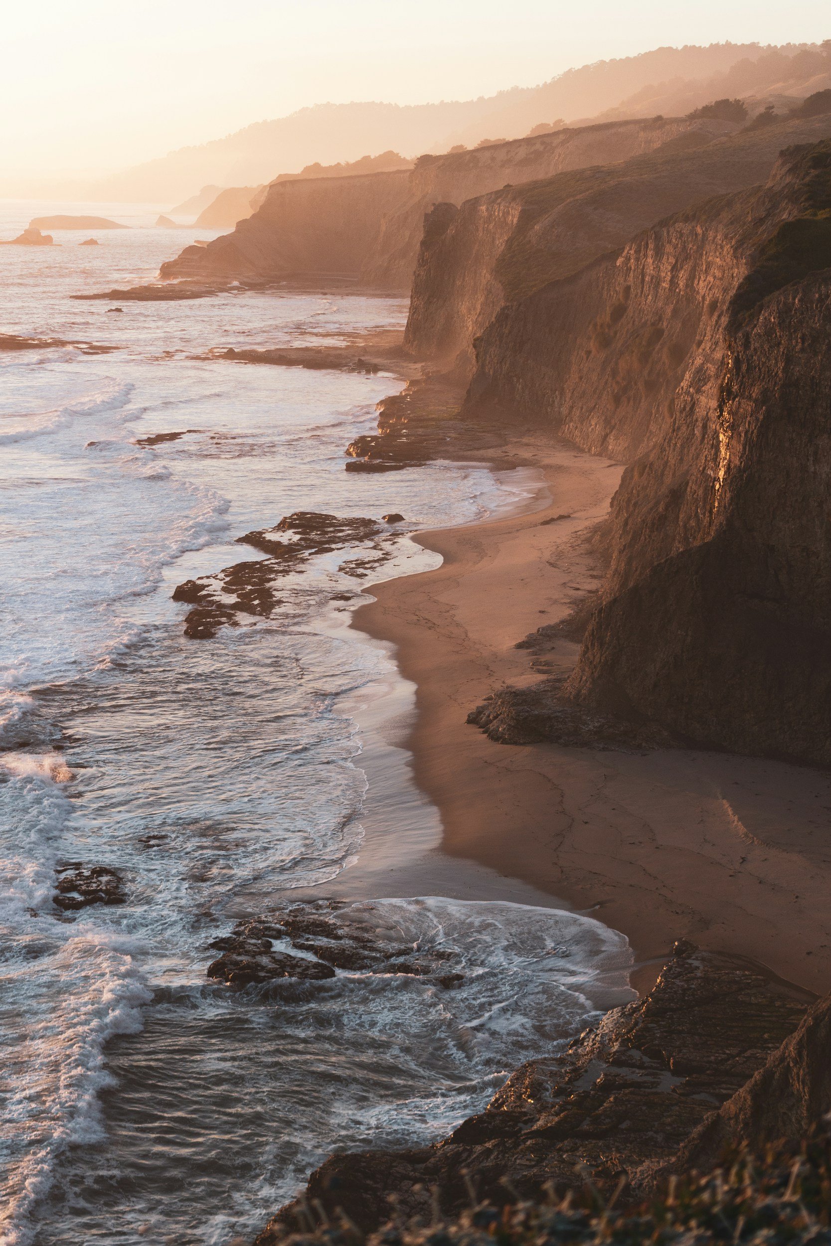 Sunset over a rugged coastline with cliffs, sandy beach, and ocean waves.