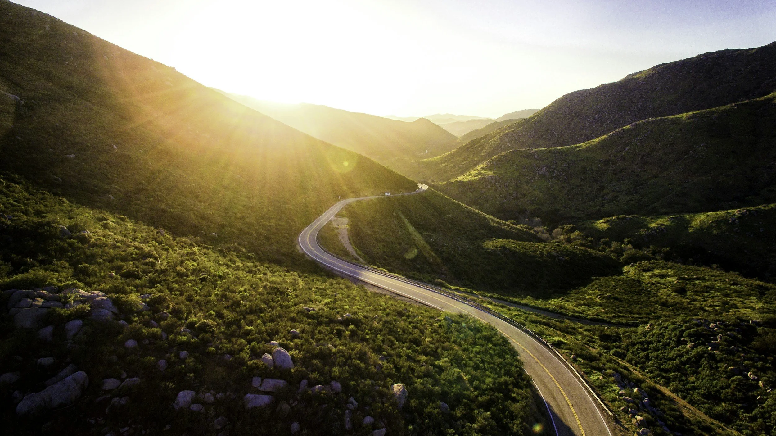 Winding mountain road in a green hilly landscape with the sun low in the sky, casting a warm glow.