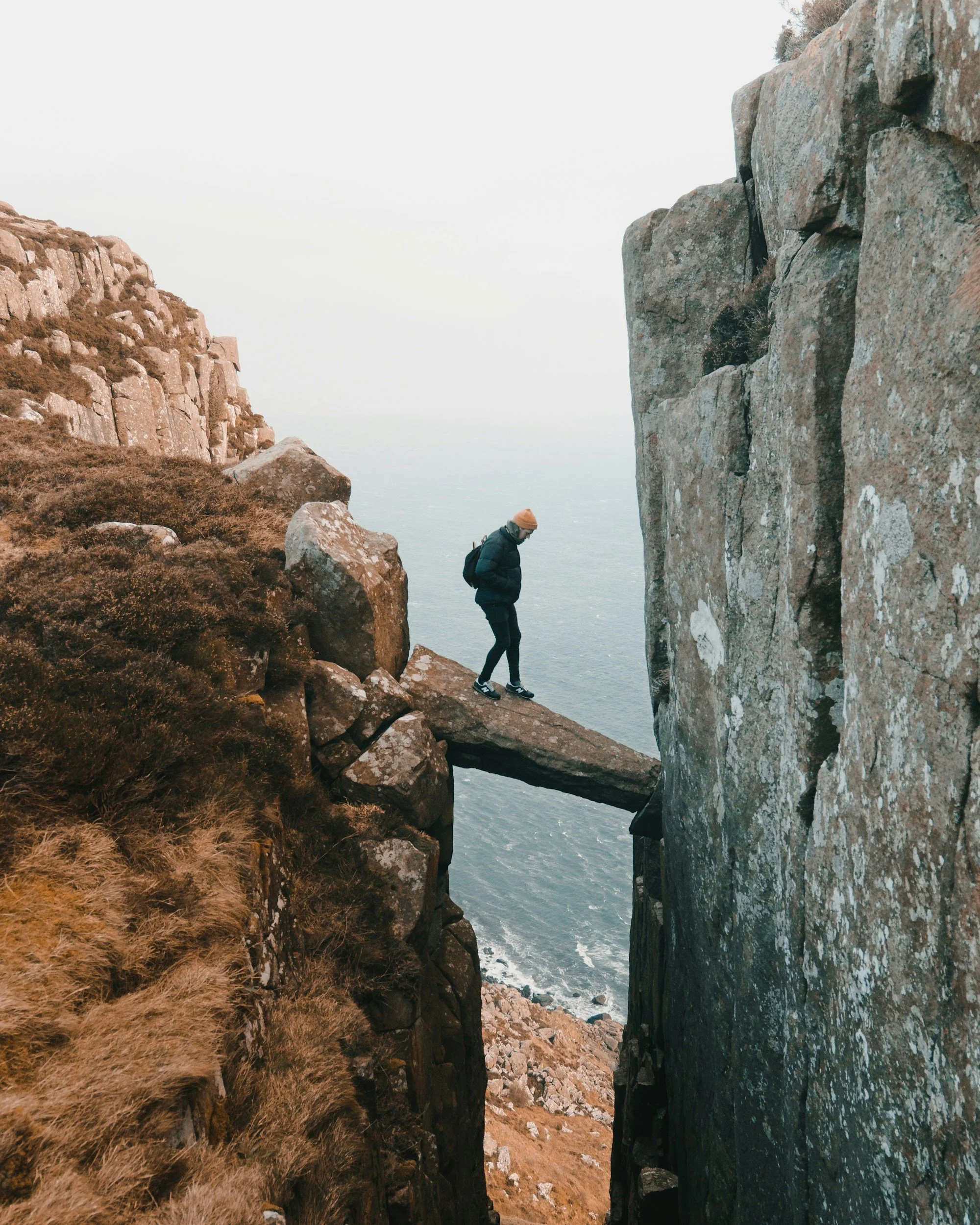 Person in dark clothing and orange beanie standing on a narrow rock ledge between two cliffs over the ocean.