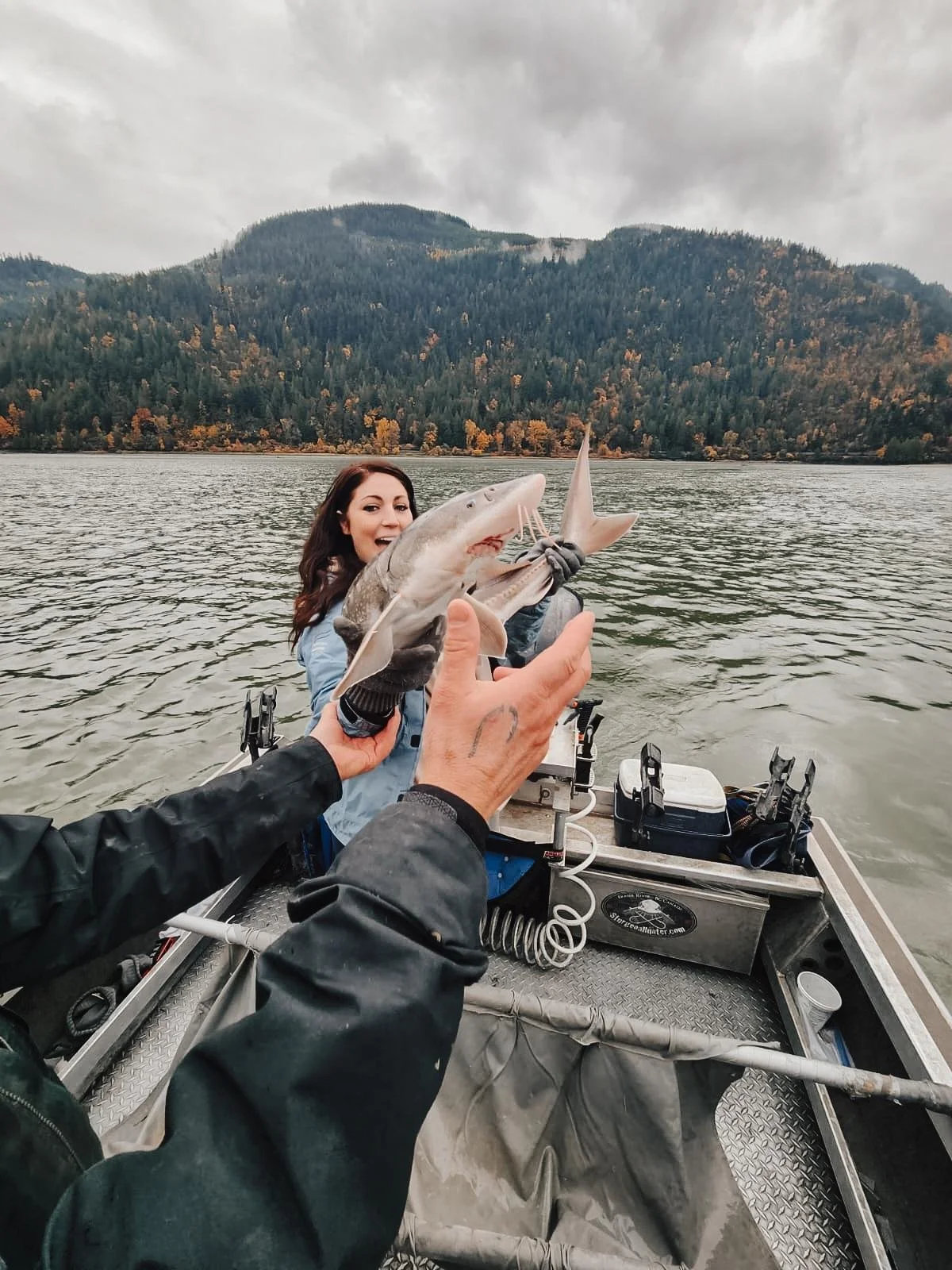 Person holding a small shark on a boat, with a woman smiling in the background and mountains with trees in the distance.