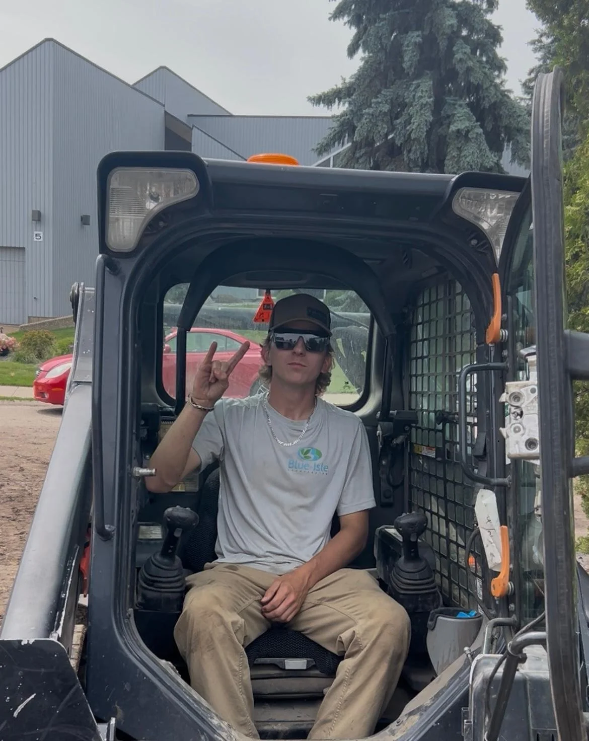 A young man sitting inside a construction vehicle, wearing sunglasses, a cap, a gray t-shirt, and khaki pants, making a hand gesture with his right hand, with an industrial building, a red car, and trees in the background.