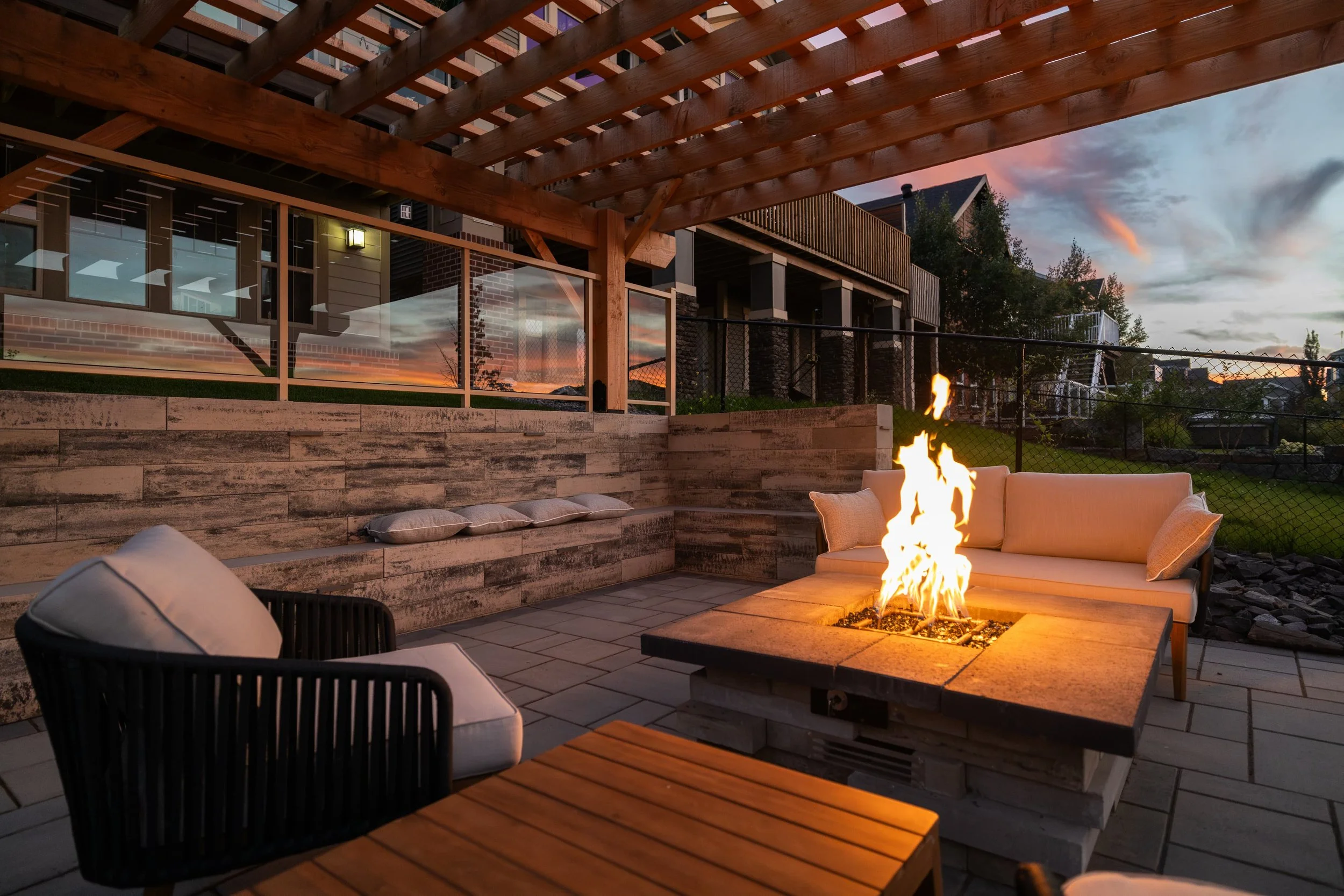 Outdoor patio with gas fireplace, seating area with cushions, wooden trellis overhead, residential houses in the background, with a sunset sky.