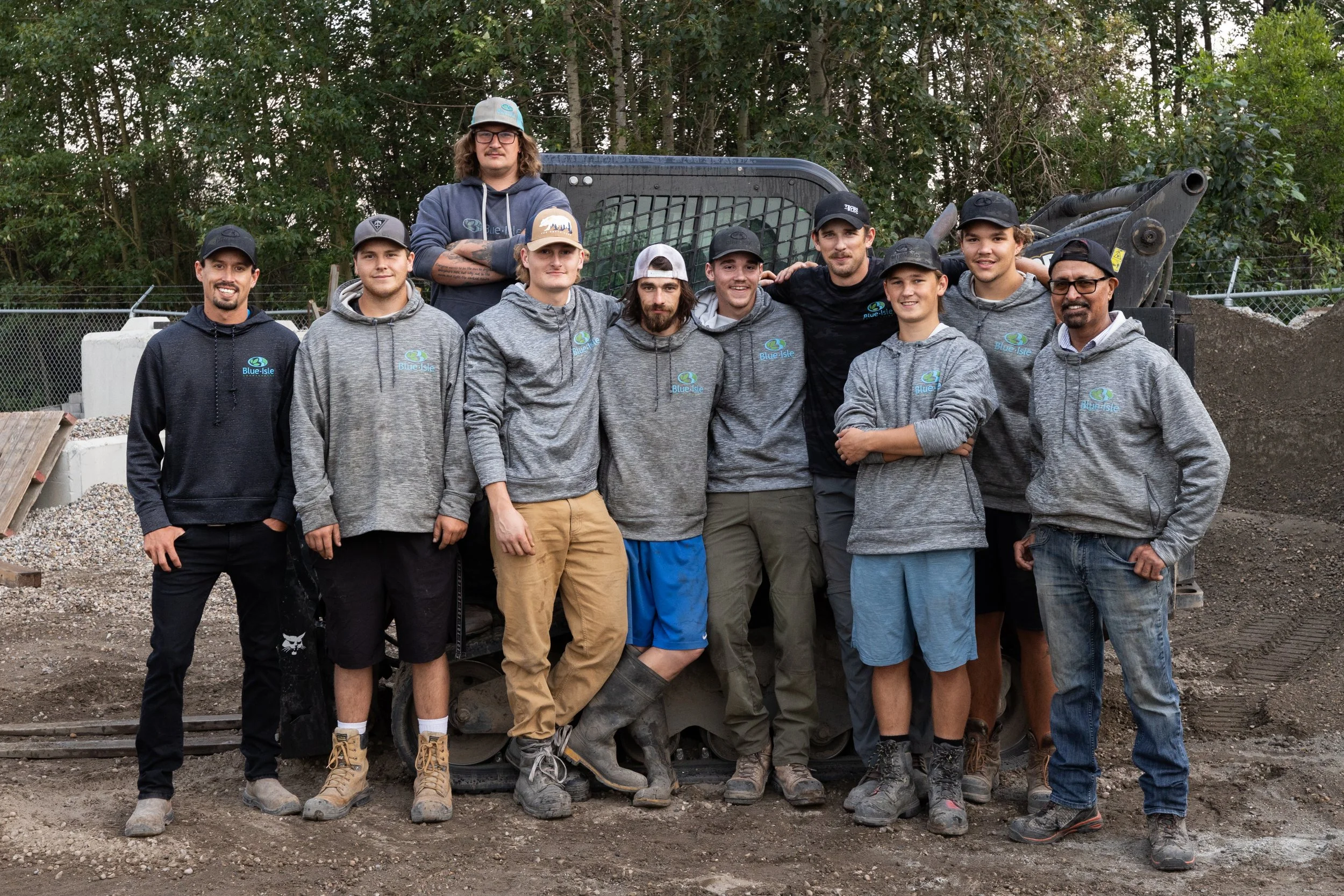 Group of ten people standing in front of construction equipment, some wearing matching gray hoodies with a blue and green logo, outdoors with trees and fencing in the background.