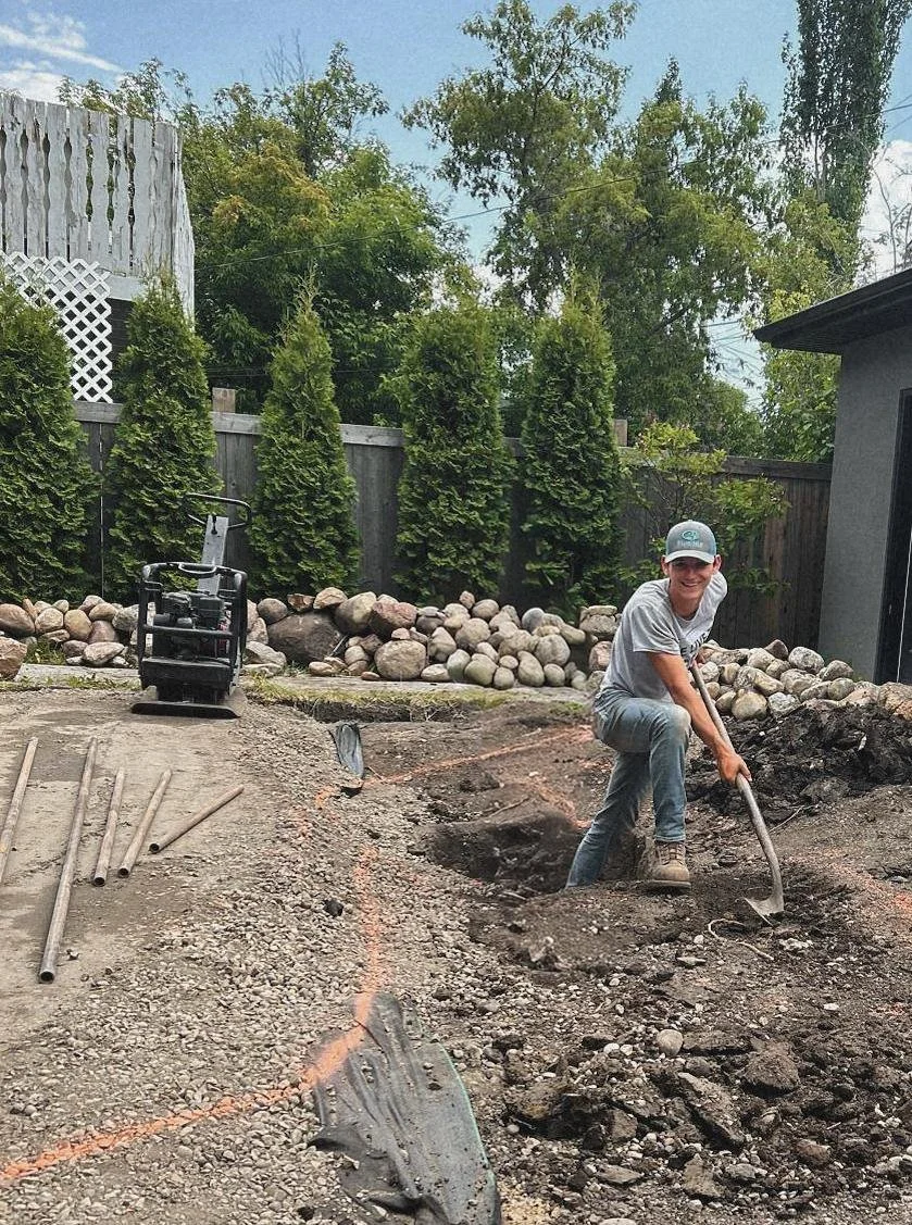 A young man is digging a trench in a backyard with a shovel, surrounded by construction tools and equipment, with tall trees and a wooden fence in the background.