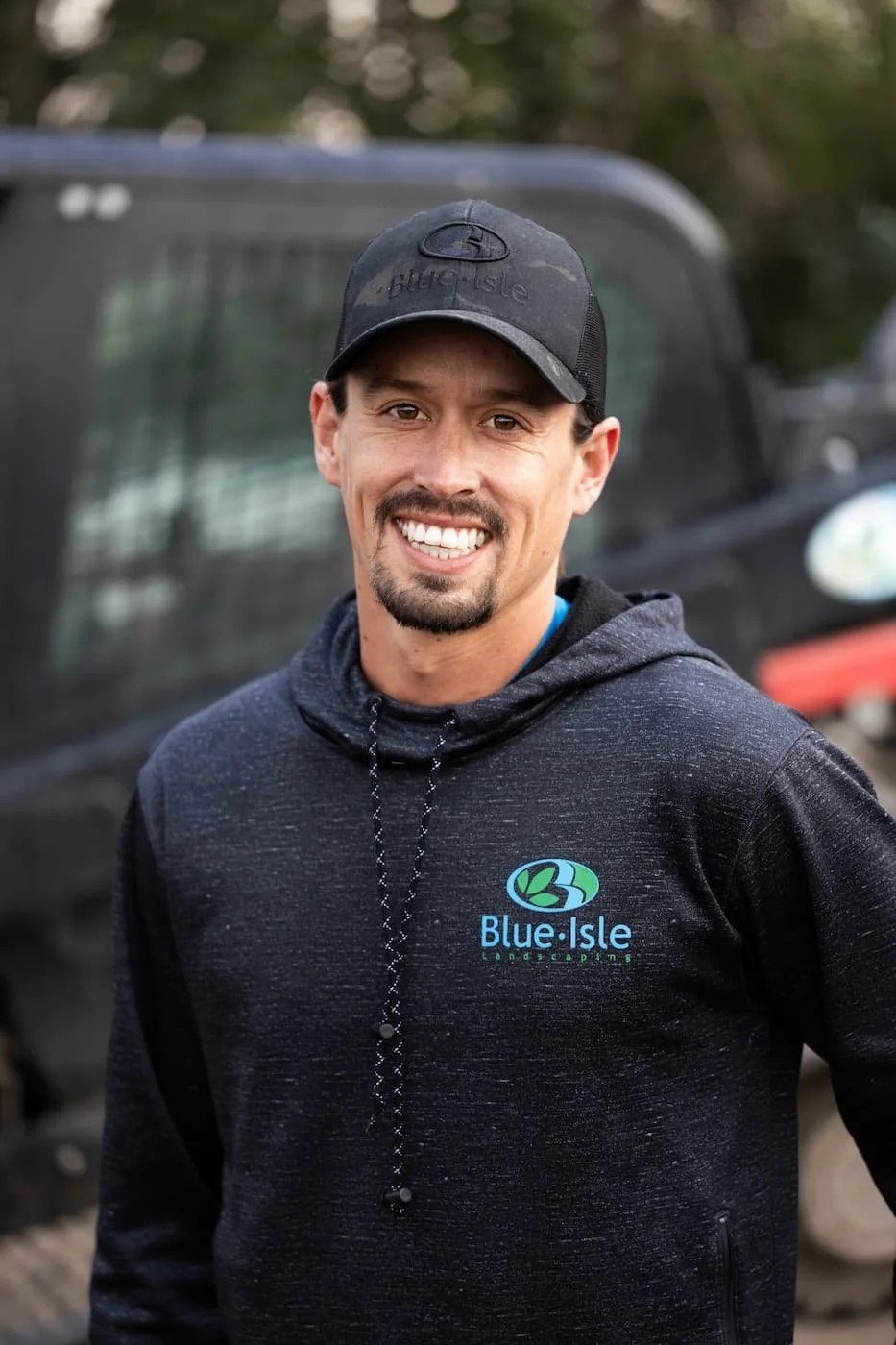 A smiling man wearing a black cap and a black hoodie with a blue-green logo that says 'Blue Isle Landscaping.' He is outdoors with a blurred view of a black vehicle in the background.