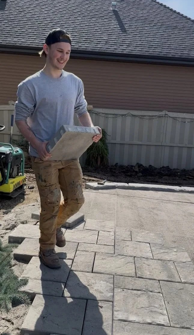 A young man in casual clothes and a backward cap working on laying stone pavers in a backyard, holding a box of paving stones.