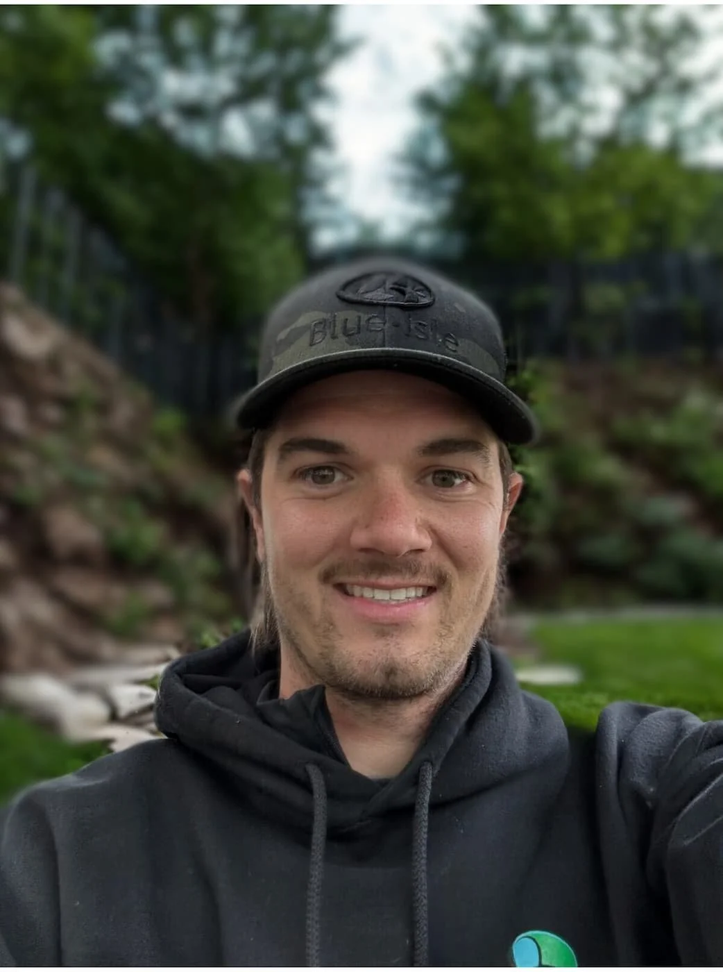 A young man smiling outdoors with trees and rocks in the background, wearing a black hoodie and a camo baseball cap.