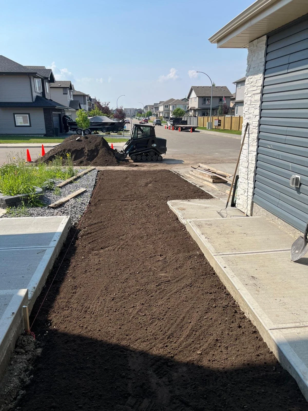 A backyard area with a newly leveled dirt path next to a house, with construction equipment such as a mini excavator and orange traffic cones in the background.