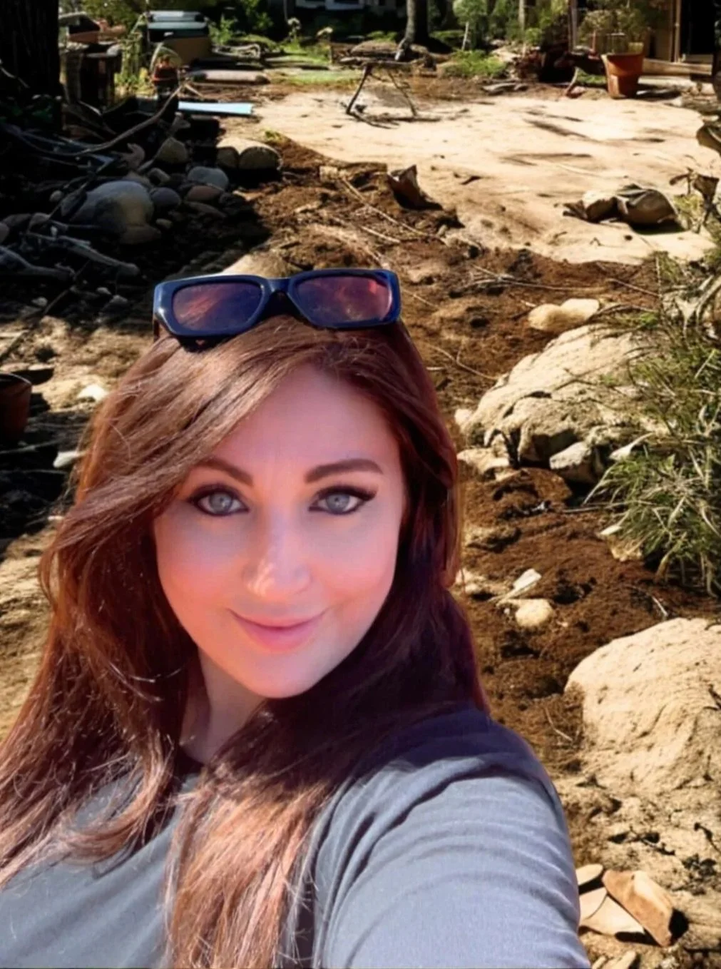 A woman with long brown hair and blue eyes taking a selfie outdoors on a sunny day, with sunglasses resting on her head. The background features a yard with dirt, rocks, some grass, and miscellaneous outdoor items.