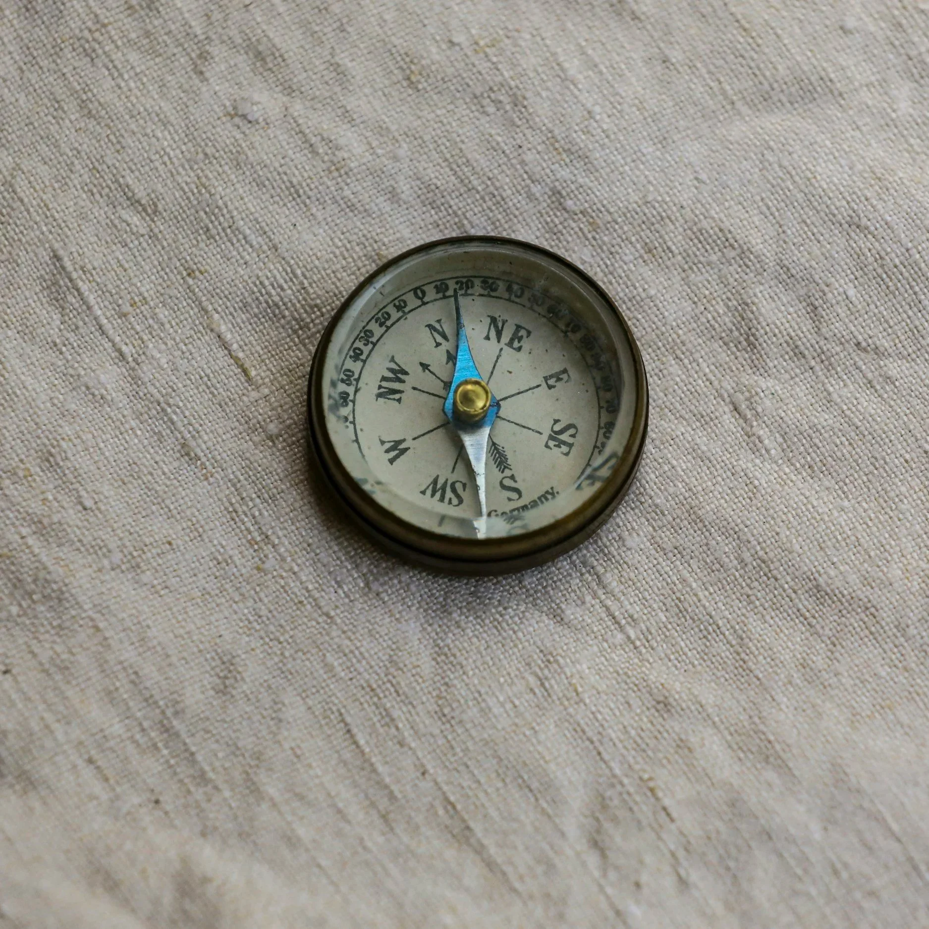 A vintage compass with a cream face and blue needle, lying on a beige fabric surface.
