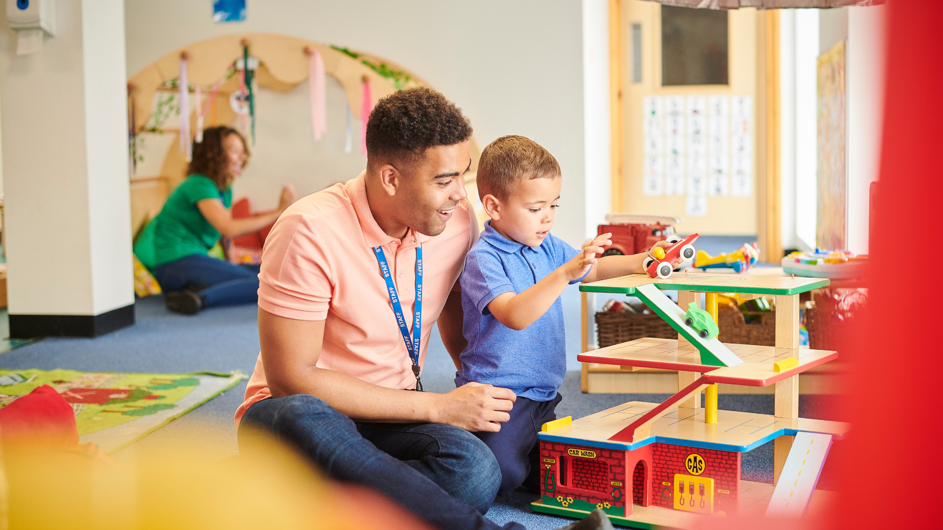 A smiling adult and a young boy playing with a colorful wooden toy garage in a playroom. A woman in green is in the background sitting on the floor.