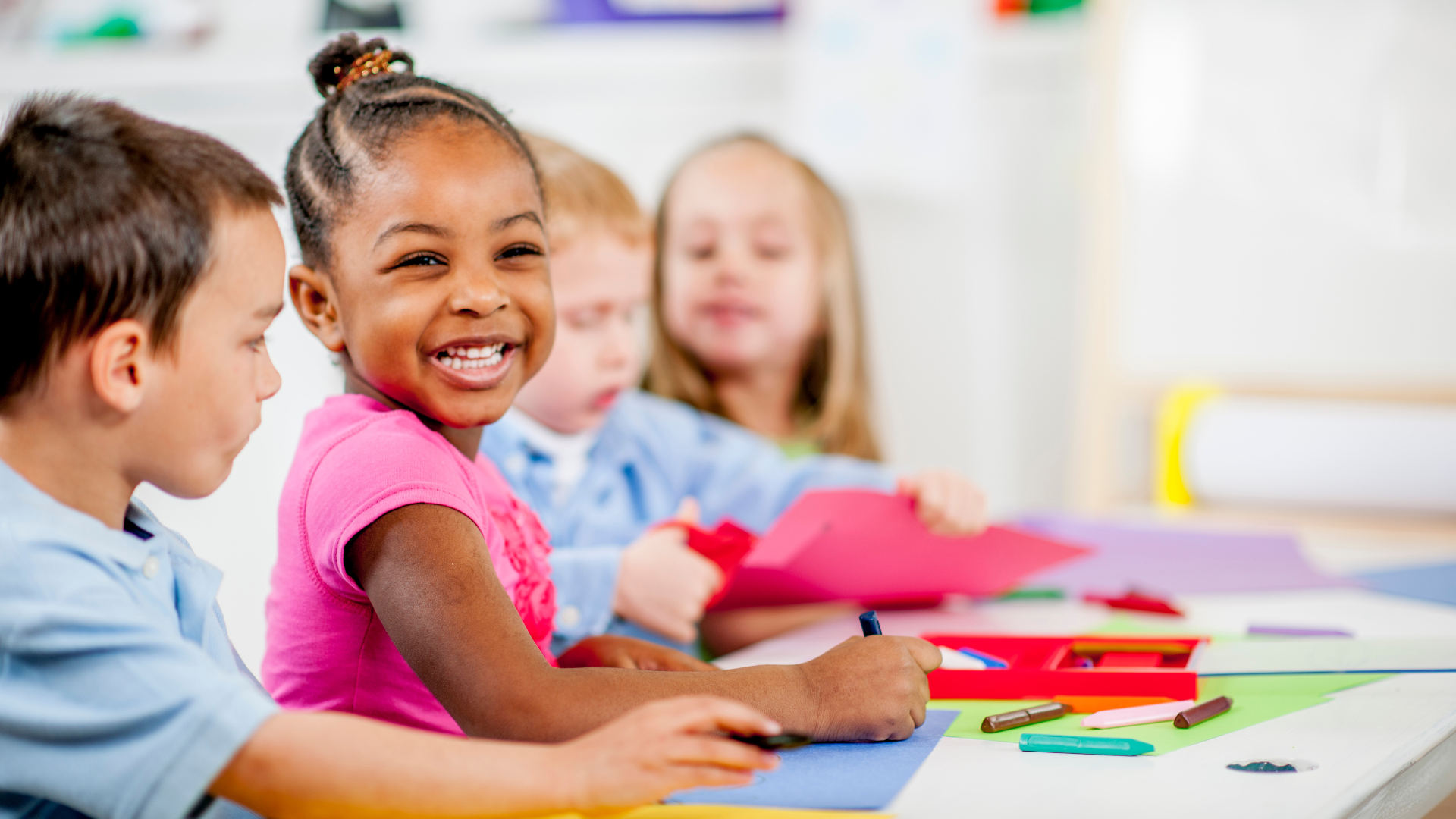 Group of young children sitting at a table engaging in arts and crafts activities, with one girl smiling at the camera.