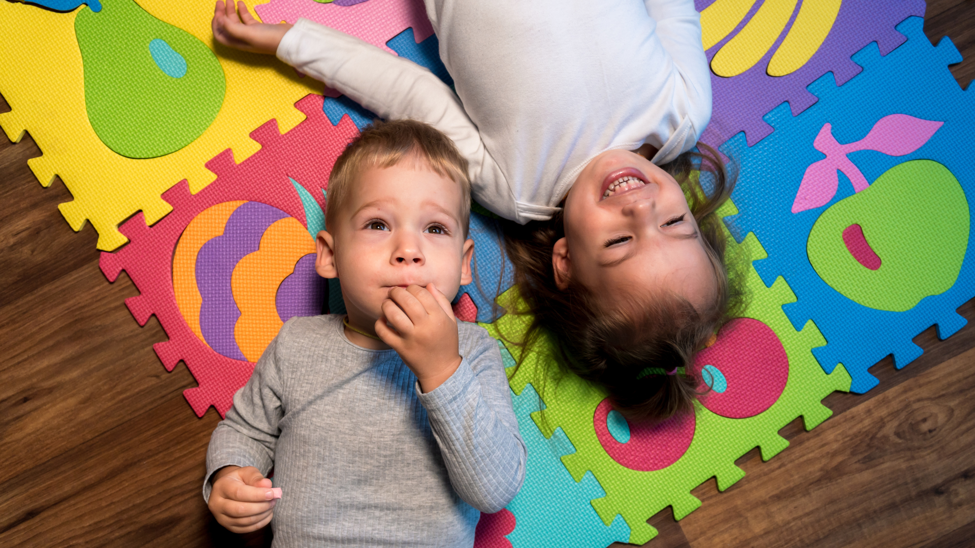Two young children, a girl and a boy, lying on colorful foam puzzle mats with fruit designs, on a wooden floor. The girl is smiling with her eyes closed, and the boy is looking thoughtful with his hand near his mouth.