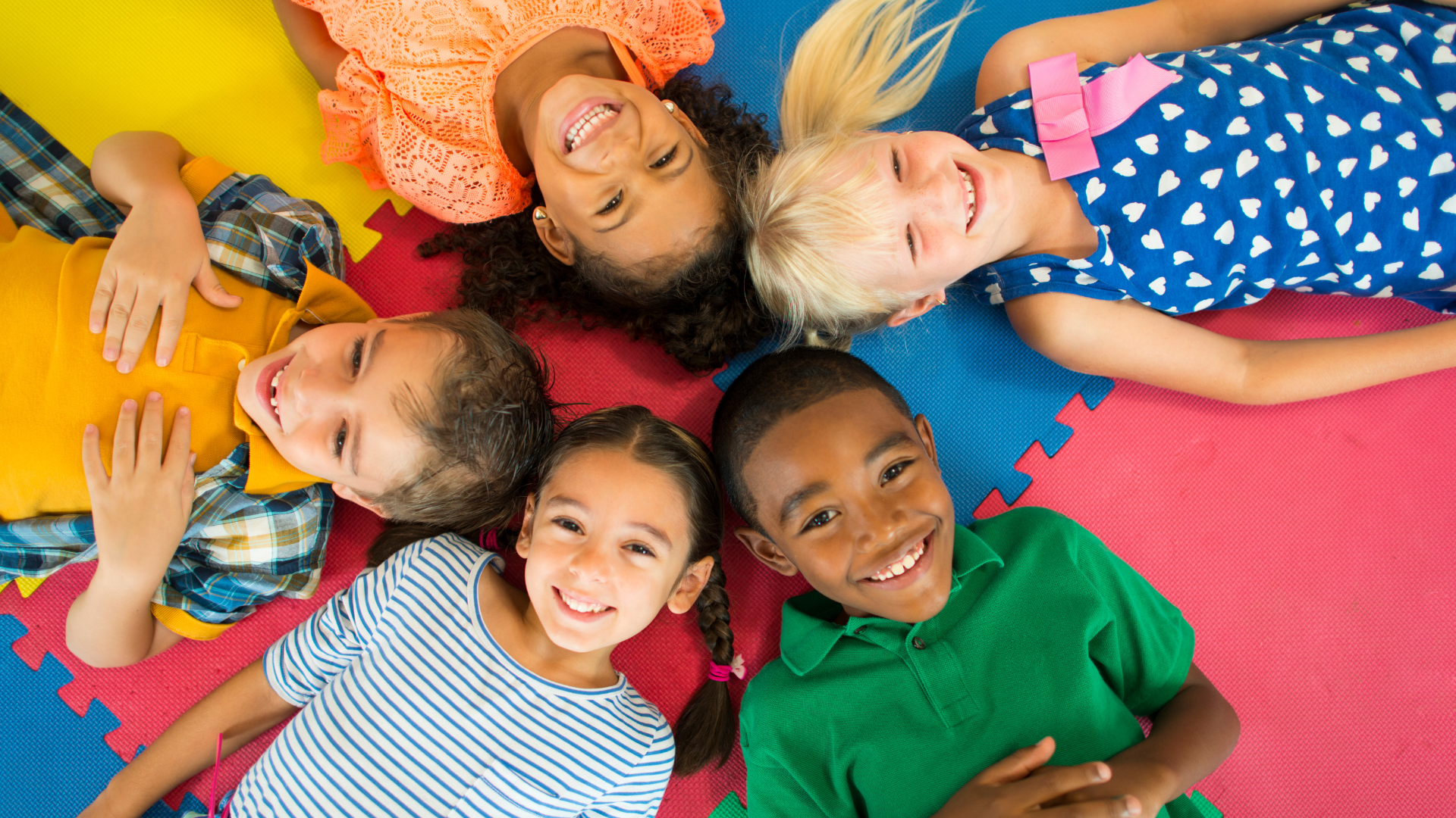 Six children lying on a colorful foam mat in a circle, smiling and looking up at the camera.