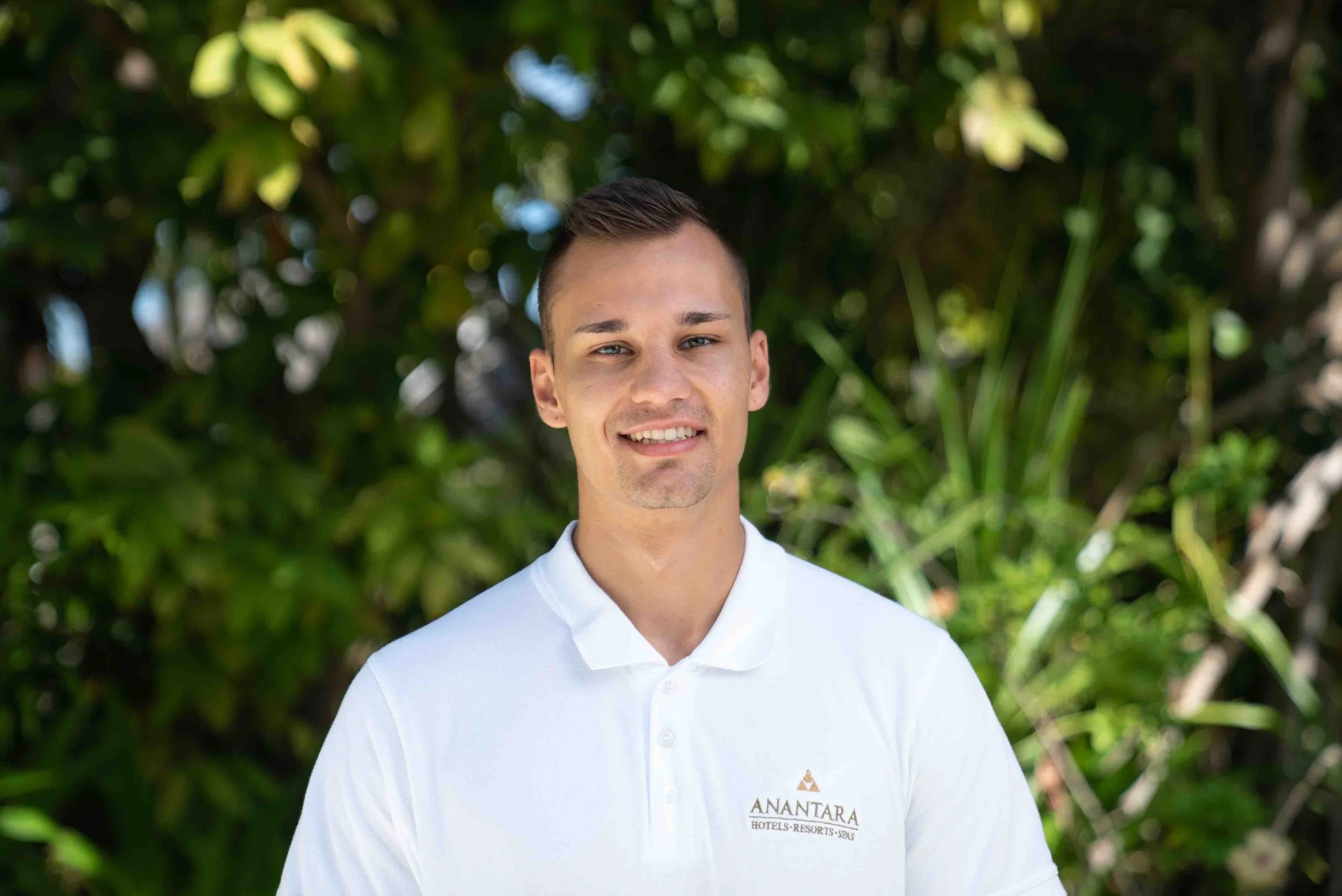 A young man with short brown hair wearing a white polo shirt with the logo "Anantara Hotels Resorts Spas" standing outdoors in front of green foliage.