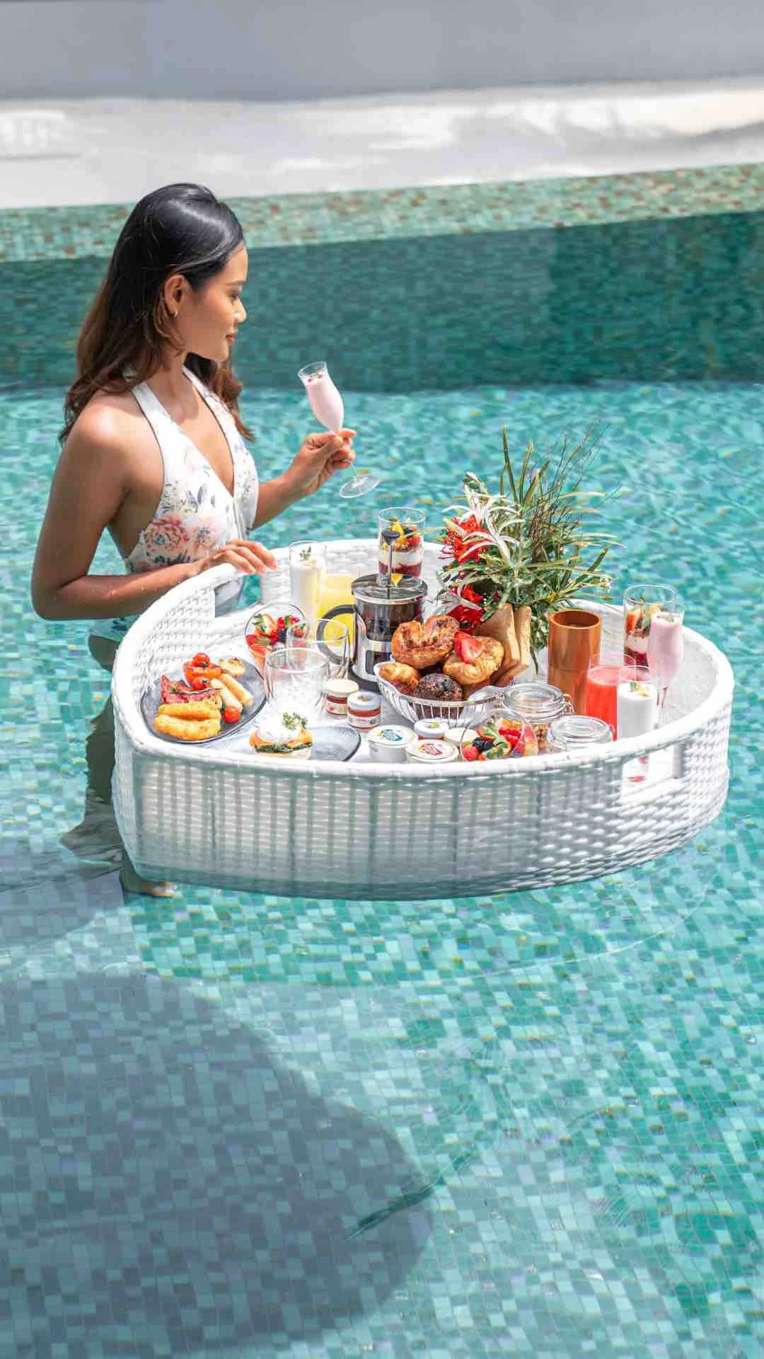 A woman standing in a swimming pool holding a glass of pink drink, with a floating breakfast tray filled with pastries, fruits, and beverages.
