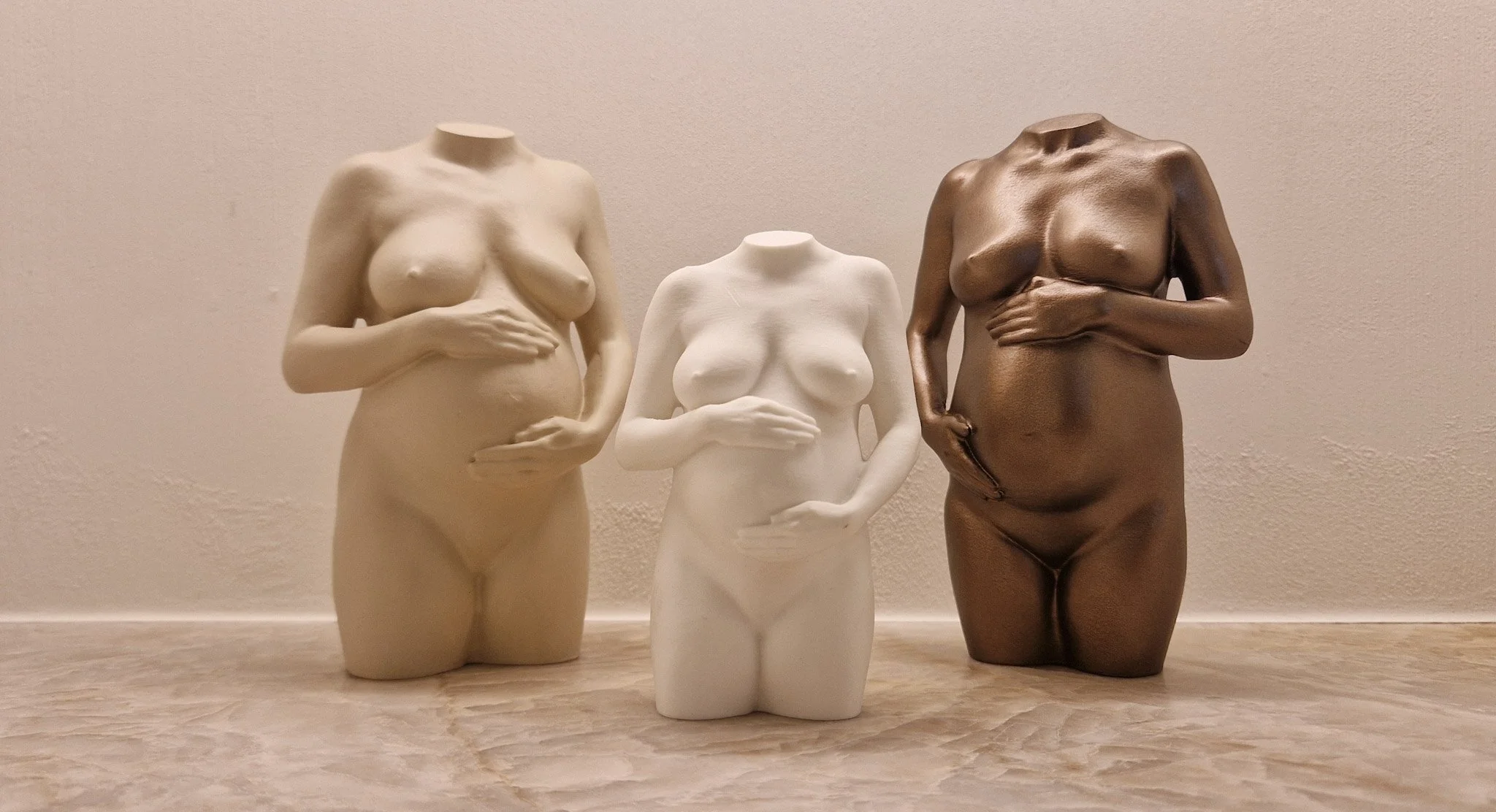 Drie sculpturen van vrouwenlichamen zonder hoofden, in beige, wit en bronskleur, geplaatst op een houten vloer tegen een effen muur.