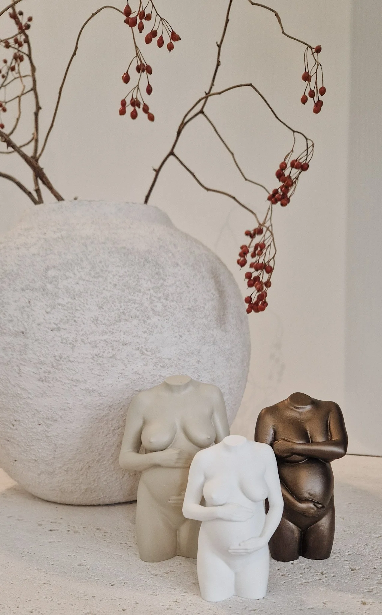 Drie naakte vrouwelijke torso beeldjes, twee in wit en één in brons, op een witte ondergrond met een grote witte keramische vaas met takken met rode bessen op de achtergrond.