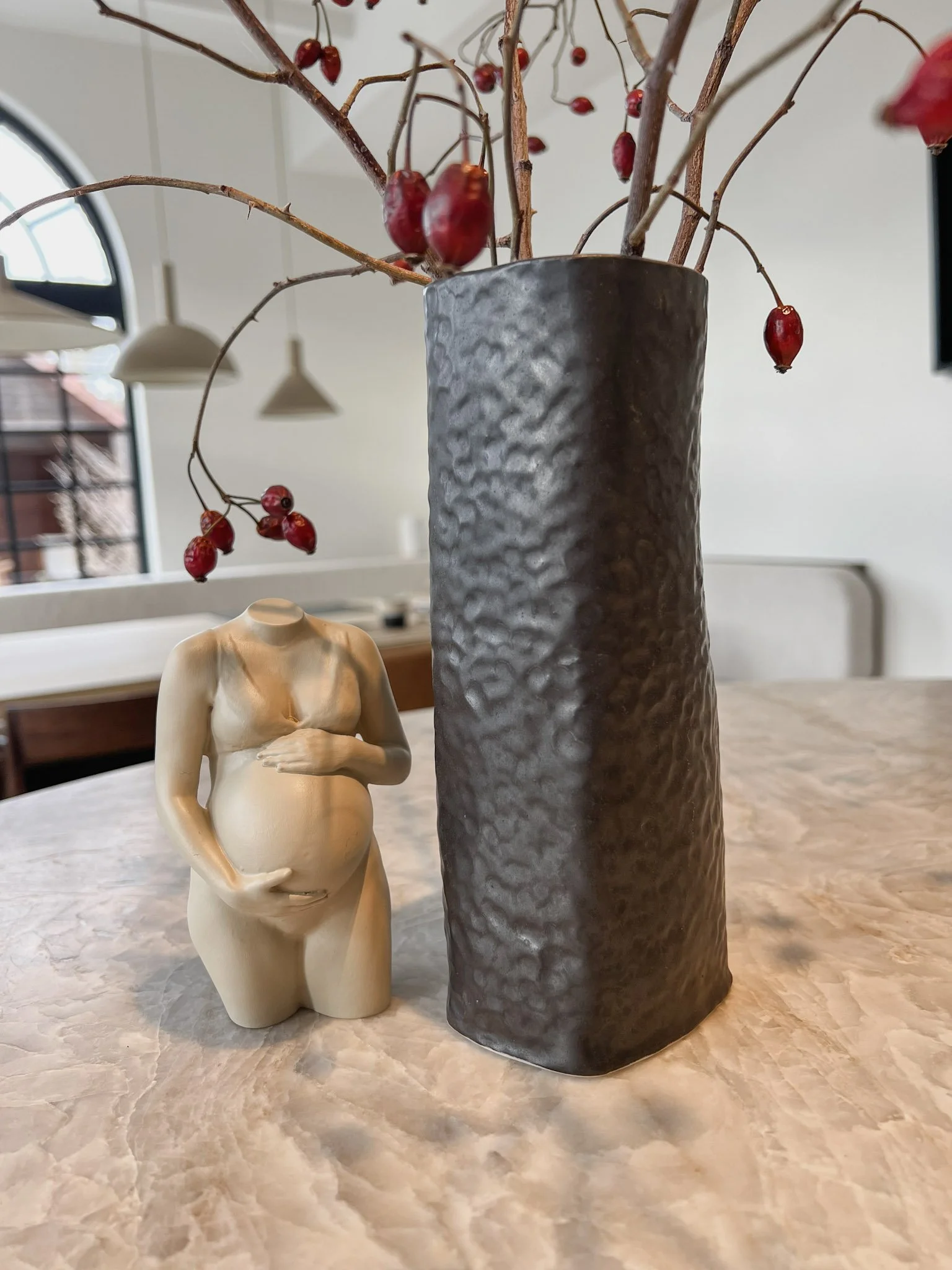 Een vaas met takken met rode bessen naast een beeld van een zwangere vrouw zonder hoofd op een tafel.