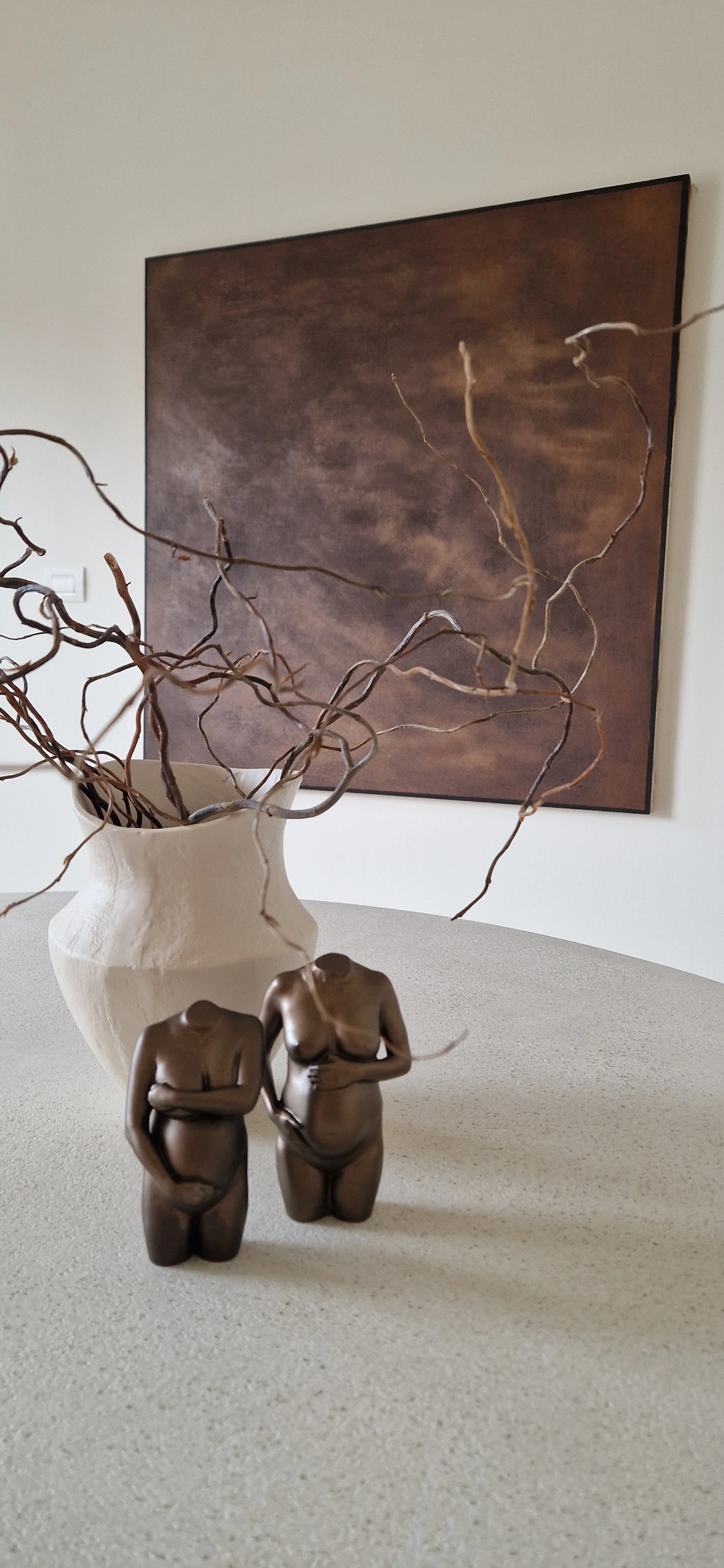 Twee bronzen beeldjes van vrouwen met handen op buik, op een tafel met een witte keramieken vaas met gedroogde takken, op de achtergrond een abstract schilderij met bruine tinten.