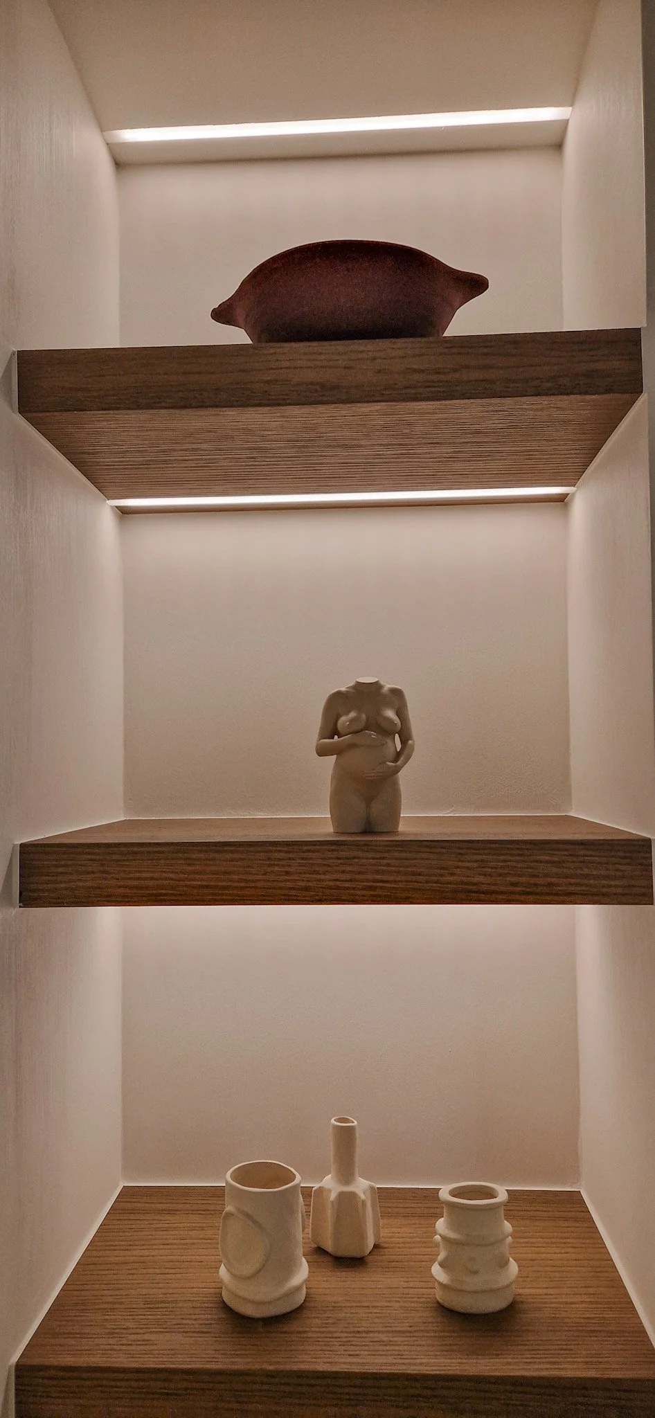 Open houten boekenplank met drie niveaus, met keramische kunstobjecten en beeldjes, met zachte binnenverlichting.