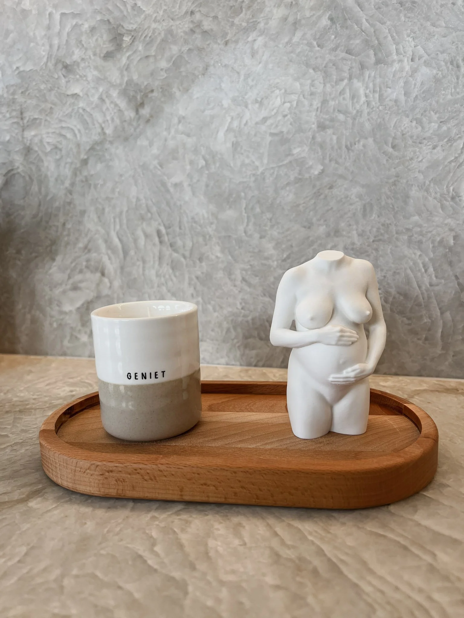 Een houten dienblad met een witte beker met de tekst 'GENIET' en een sculptuur van een naakte vrouw op een beige tafel met een grijze muur op de achtergrond.
