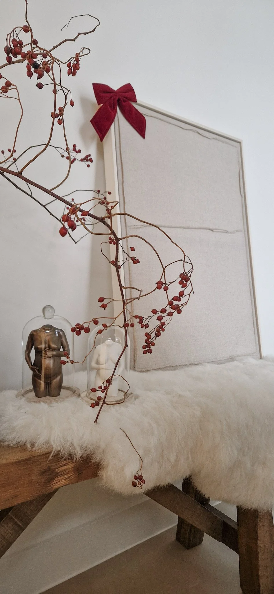 Decoratief herfst- of winterstuk met takken, rode bessen, een glazen beeldhouwwerk van een half-naakte vrouw, een witte kaars, en een wit prikbord met rode strikopmaak, geplaatst op een houten tafel met een lichte, zachte pluche kleed.