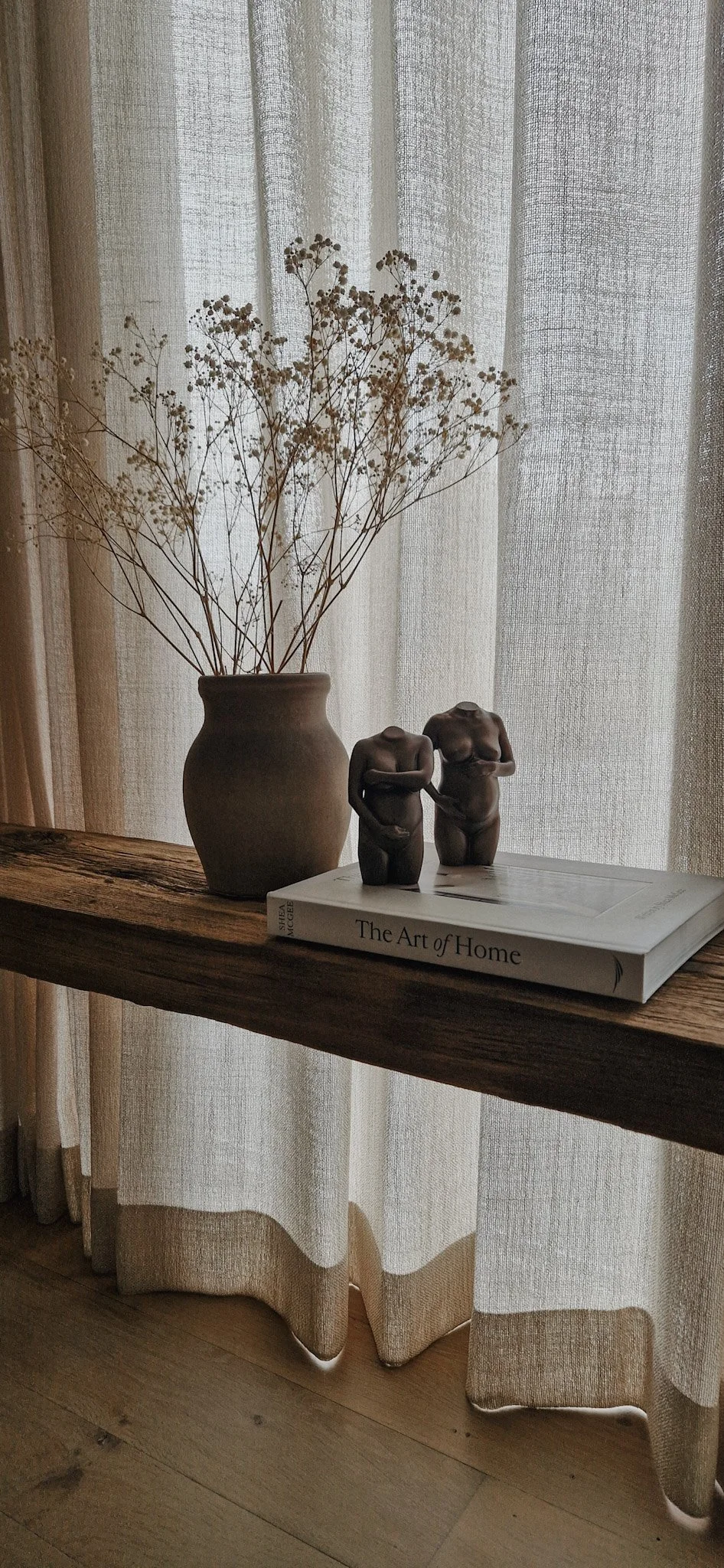 Een boek met de titel 'The Art of Home' ligt op een houten plank, naast twee statuetten van naakte vrouwen met armen over de buik, en een vaas met gedroogde witte bloemen, voor een raam met lichte gordijnen.