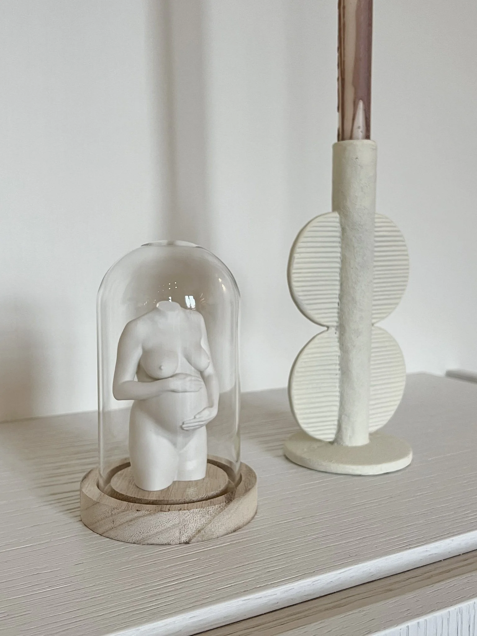 Decoratieve sculptuur van een naakte vrouw onder een glazen stolp op een houten basis, naast een abstract beeld van horizontale lijnen en een verticale staaf op een plank.