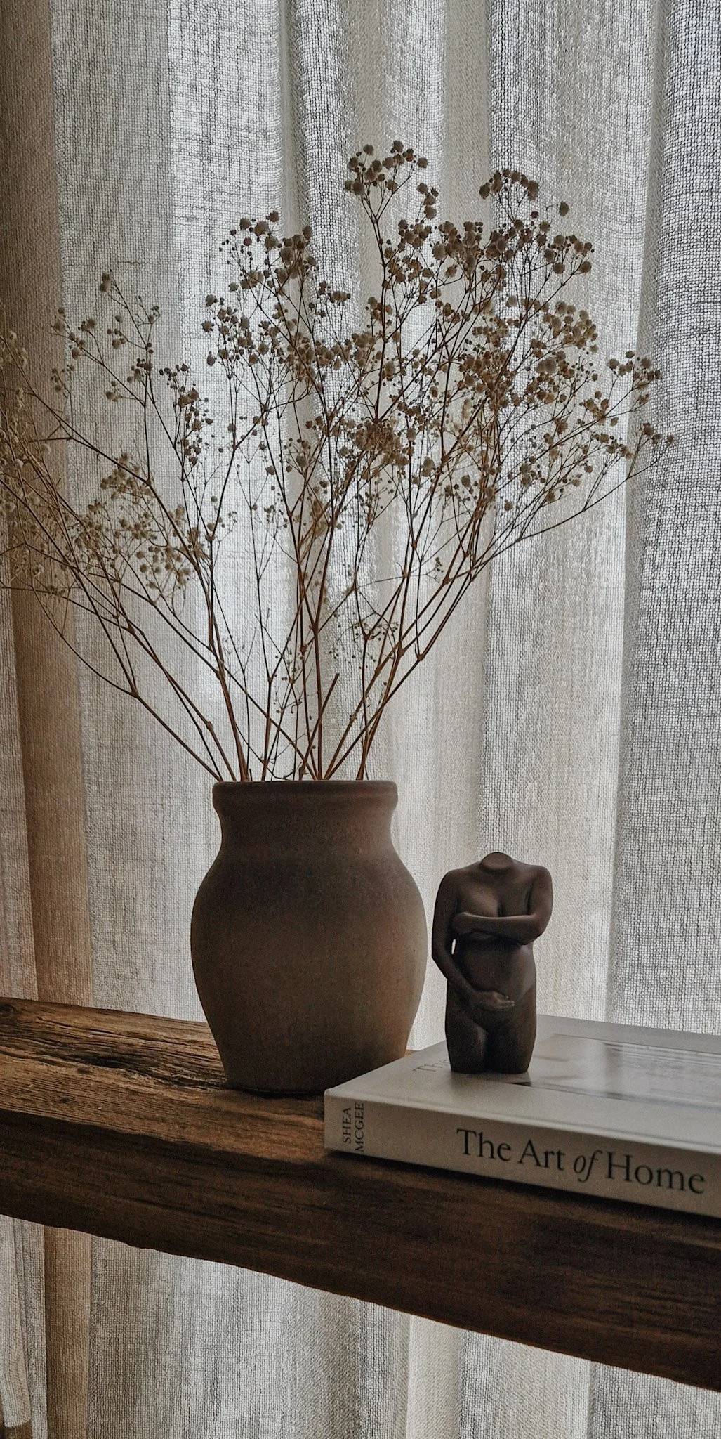 Een vaas met gedroogde witte bloemen, een sculptuur van een naakte persoon met armen over de buik, en een boek getiteld 'The Art of Home' op een houten plank voor een beige gordijn.