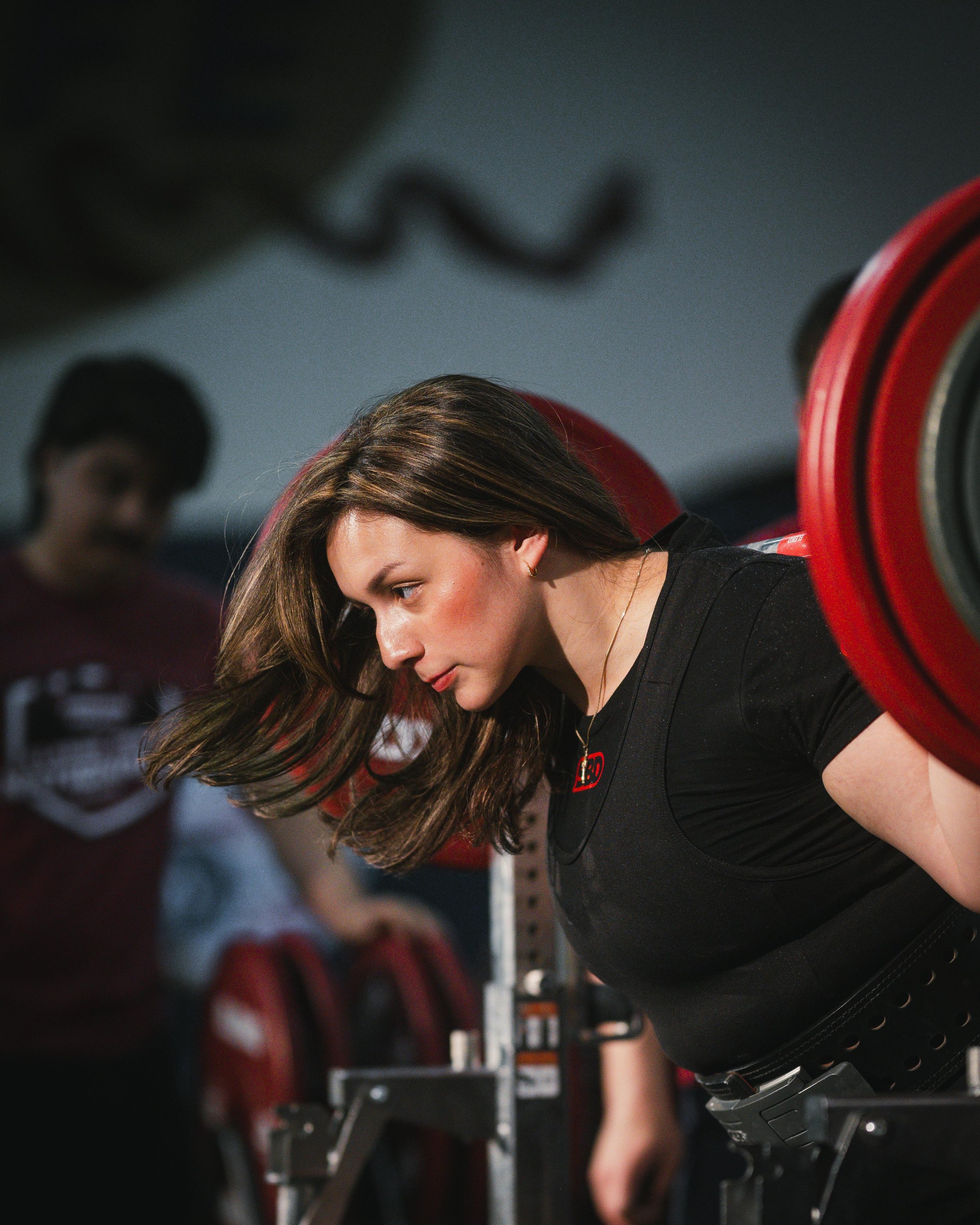 A woman with long brown hair and a serious expression is preparing to lift a barbell loaded with red weight plates at a gym, with a man blurred in the background.