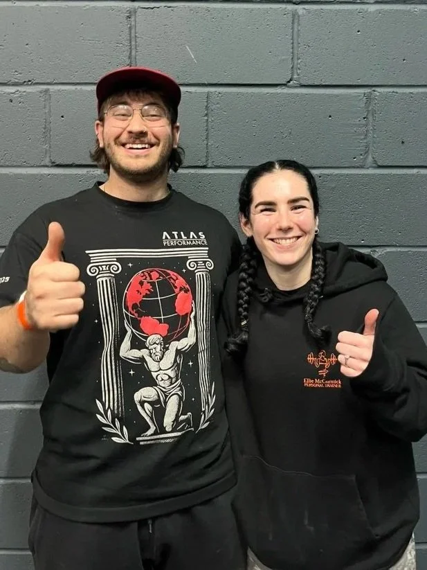 Two smiling people standing against a gray brick wall, giving thumbs up. The person on the left is a young man with glasses, a red cap, and a black T-shirt with a graphic design. The person on the right is a young woman with braided hair and a black hoodie.