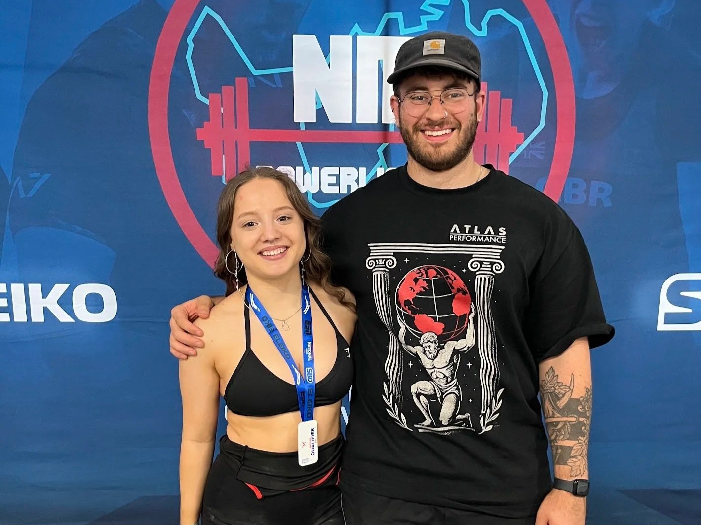 A smiling young woman and a smiling young man stand together in front of a backdrop with a logo. The woman wears a black sports bra and a medal around her neck. The man wears a black graphic t-shirt, a black cap, and has tattoos on his left arm. They are posing with their arms around each other, smiling at the camera.