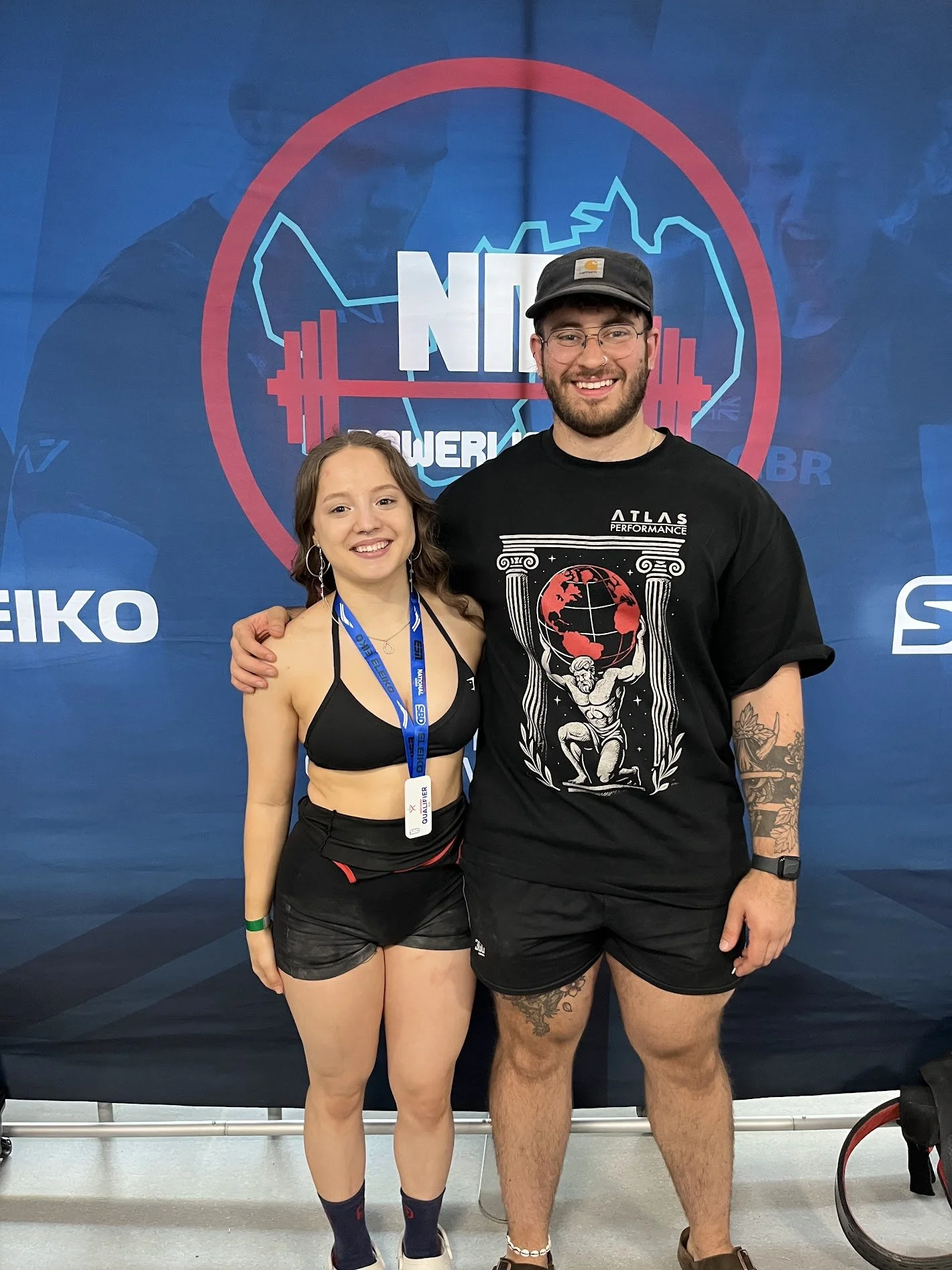 A young woman and a young man pose together, smiling, in front of a backdrop with a logo that reads 'NP' and features a barbell and abstract city skyline. The young woman is wearing a black sports bra, black shorts, and a blue lanyard. The young man is wearing a black t-shirt with a graphic print and black shorts, and has tattoos and a watch on his left wrist.