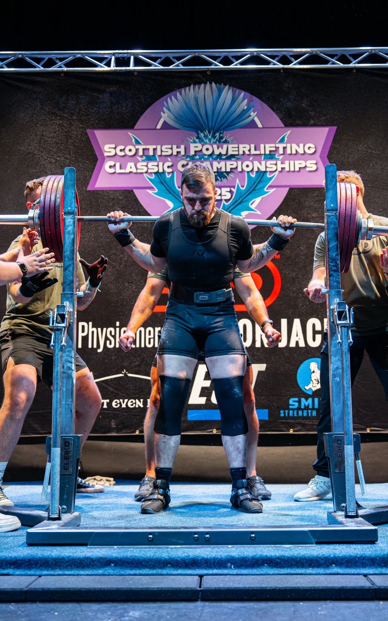 Powerlifter performing a squat at the Scottish Powerlifting Classic Championships, with spotters on each side and a banner in the background.