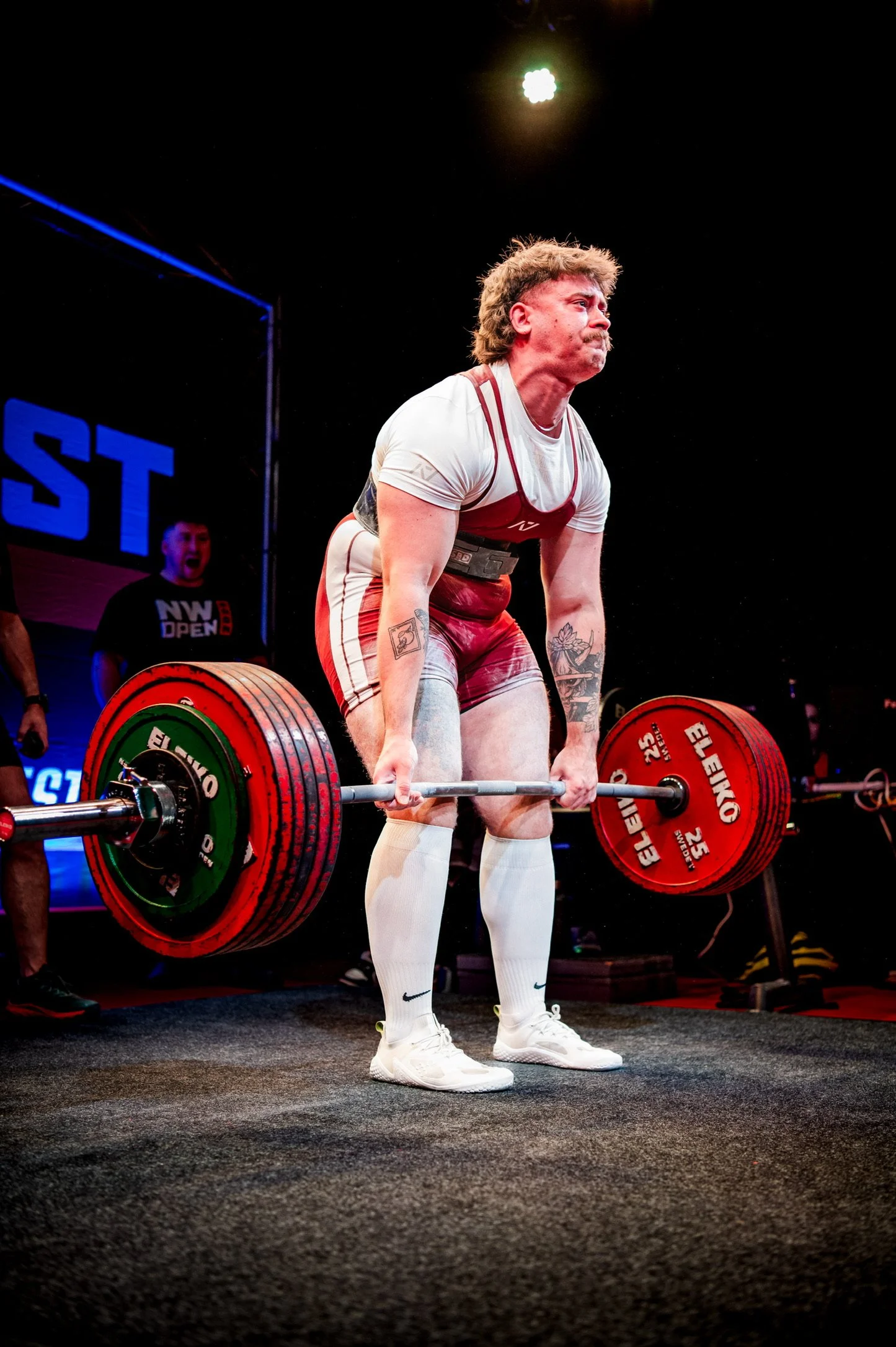 A powerlifter in a white shirt and red shorts deadlifting a loaded barbell during a competition.