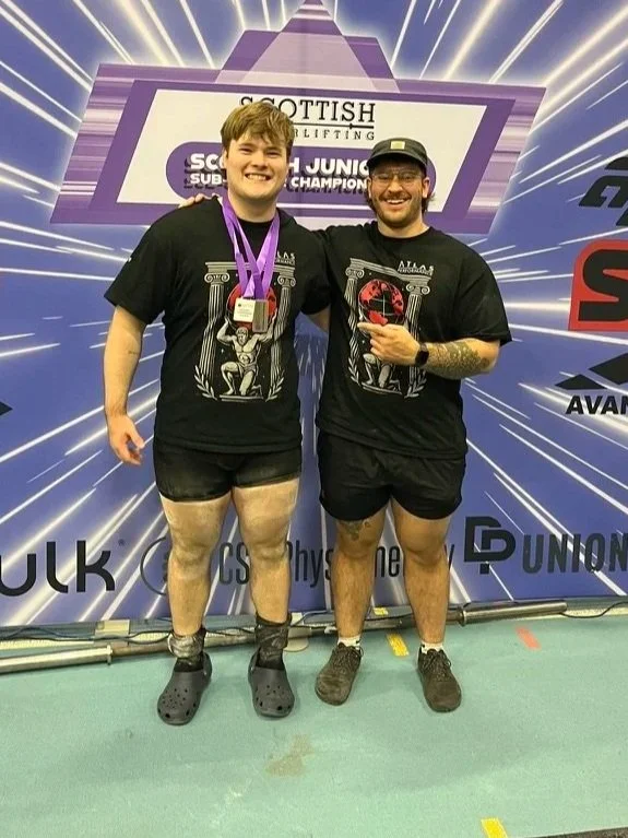 Two male powerlifters standing side by side, smiling, with medals hanging around their necks. They are dressed in black T-shirts with a graphic design, and black shorts. Their legs are shaved, and they are wearing different footwear—one with Crocs and the other with sneakers. Behind them is a backdrop with logos and the words 'Scottish Powerlifting' and 'Scottish Junior Champion,' indicating a competition setting.